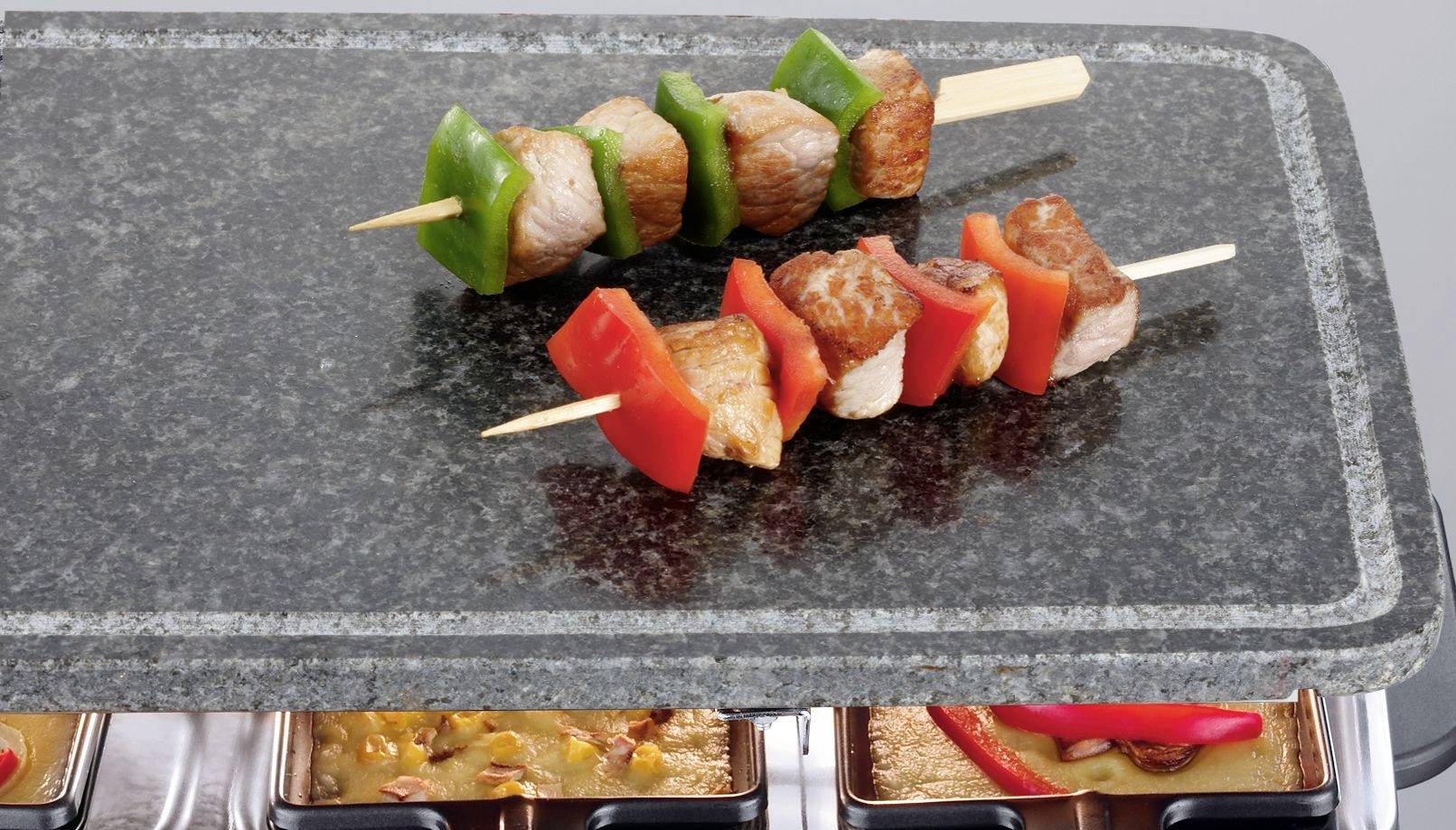 Deux brochettes de viande avec des morceaux de poivrons verts et rouges reposent sur une pierre chaude. En dessous, des petits plats à raclette sont placés dans un appareil à raclette.