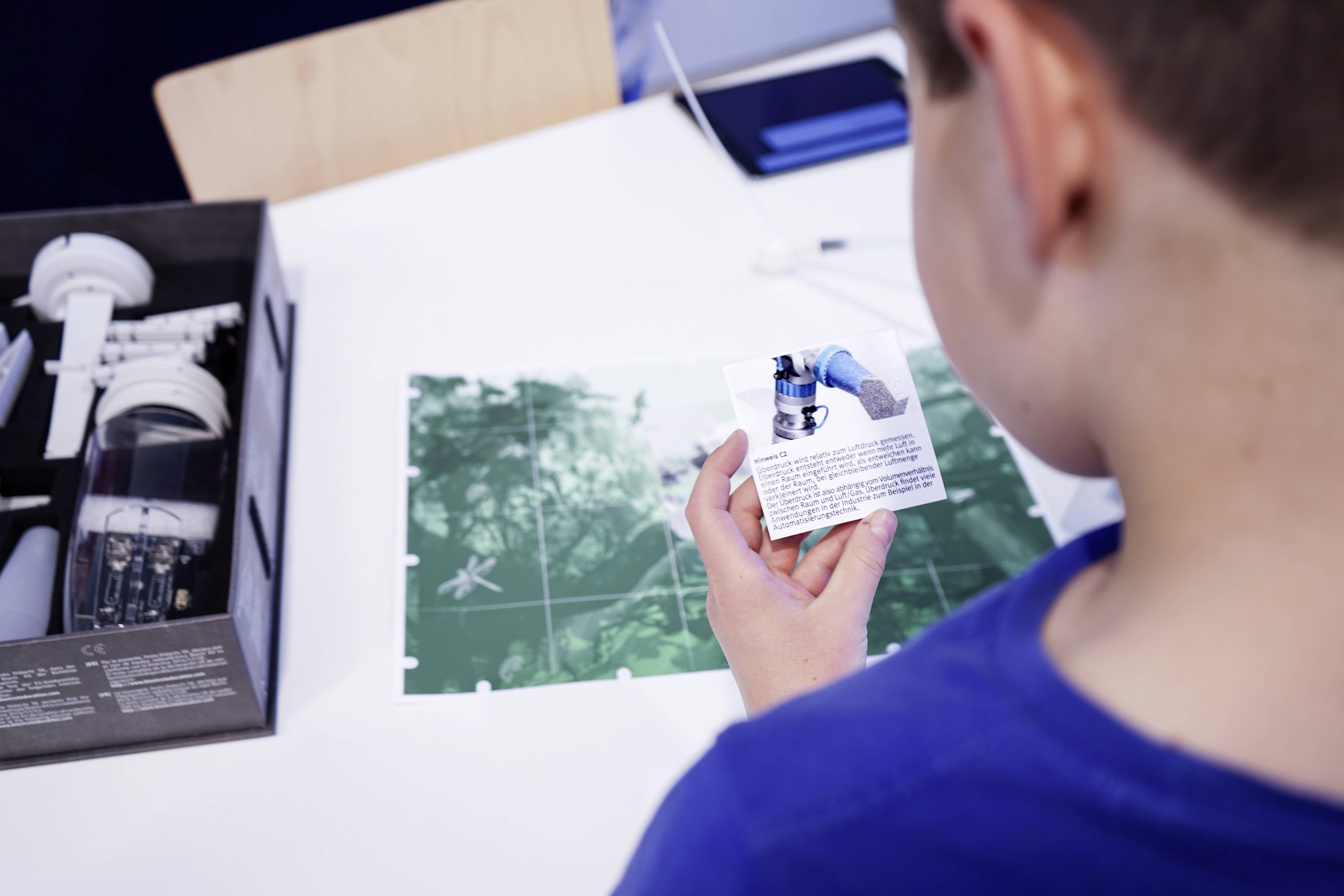 Un enfant portant un t-shirt bleu examine une carte. Sur la table, il y a un carton ouvert contenant un bras robotique jouet.