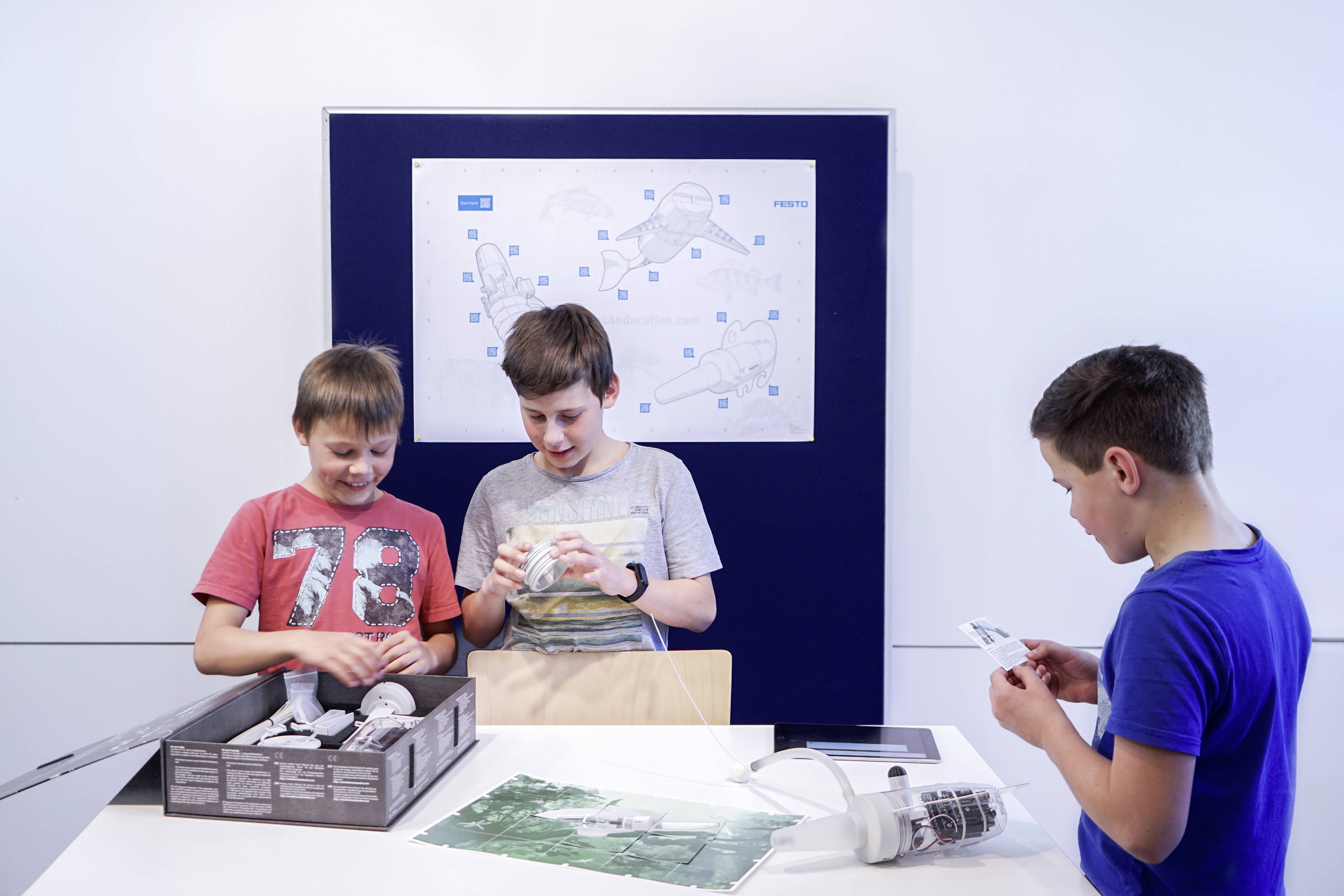 Trois garçons sont assis à une table et travaillent sur un kit de robot. En arrière-plan, un plan de construction est accroché au mur.