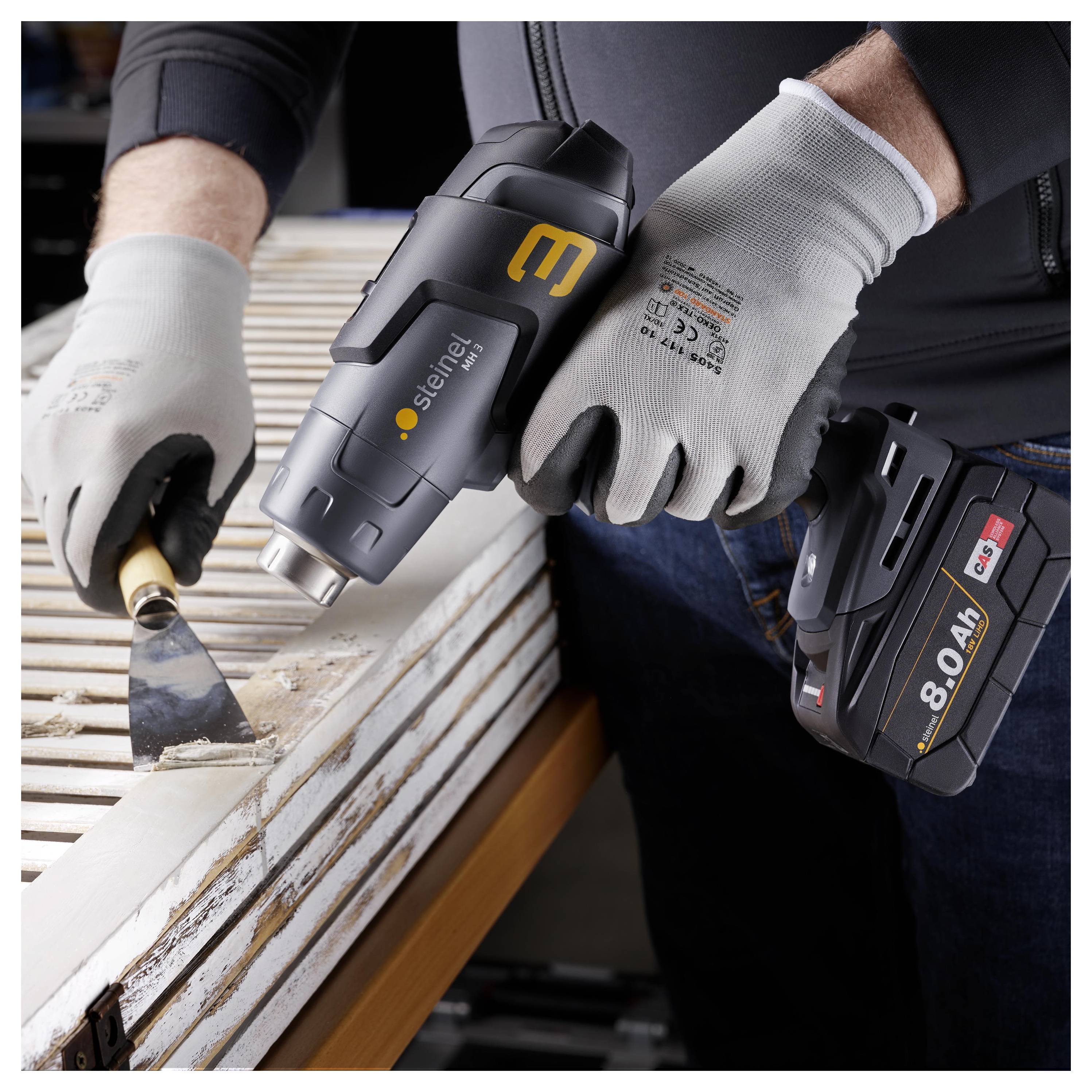 Personne portant des gants utilisant un décapeur thermique sans fil Steinel pour enlever la peinture d'une surface en bois, avec un bloc-batterie attaché.