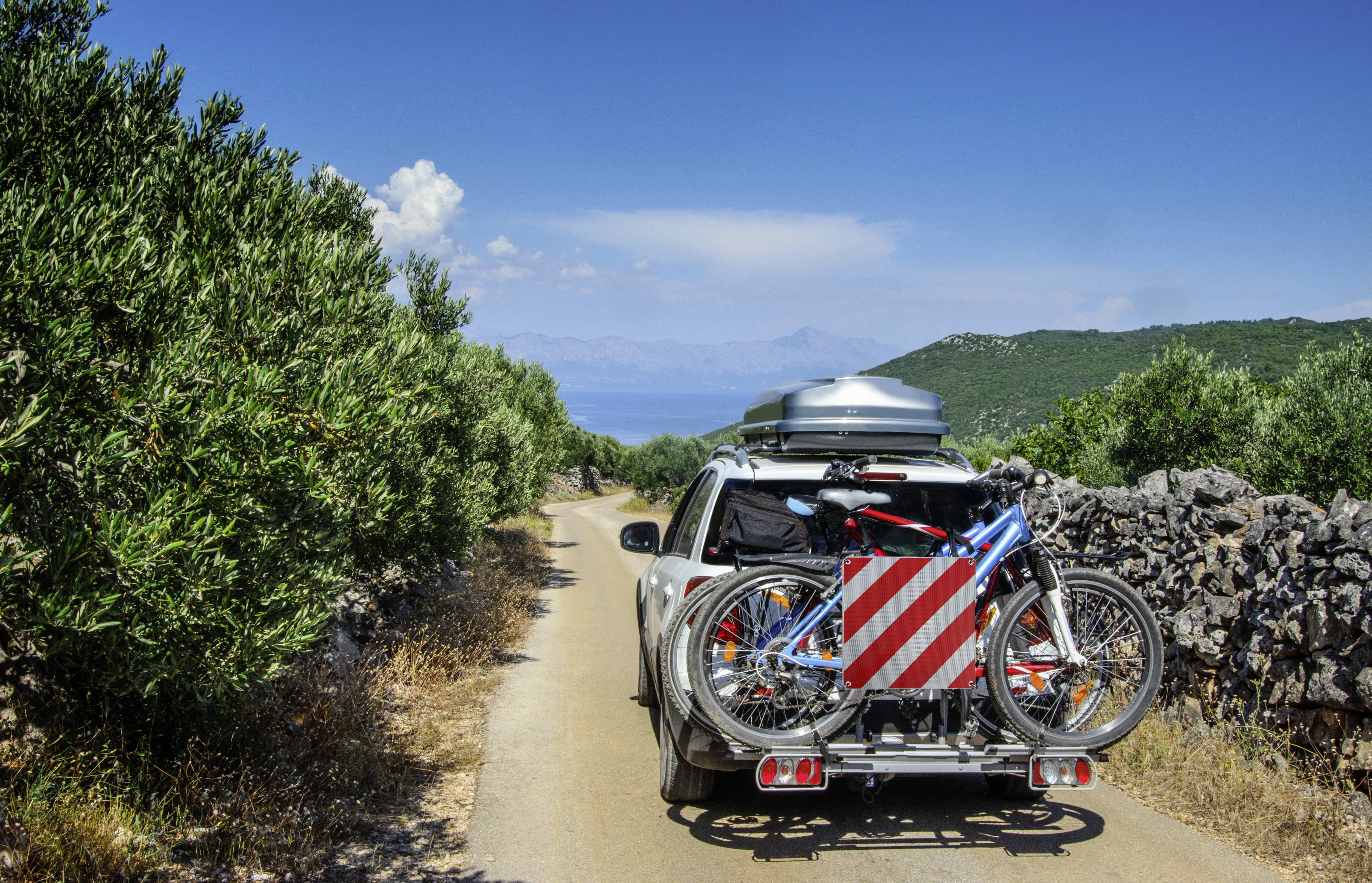 Une voiture roule sur une route étroite à travers un paysage verdoyant, avec deux vélos sur un porte-vélos arrière et un coffre de toit.
