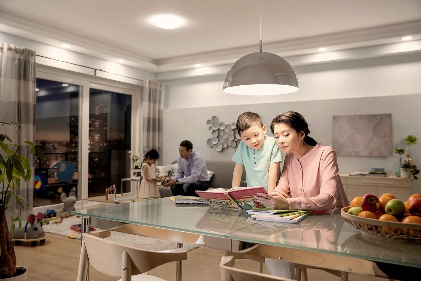 Une femme lit un livre à un garçon à une table, tandis qu'un homme joue avec une fille dans le salon en arrière-plan.