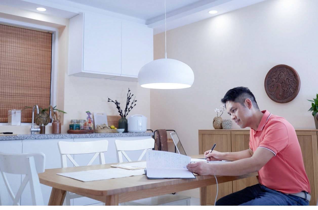 Un homme portant une chemise rouge-orangée est assis à une table de cuisine et prend des notes dans un carnet. Une cuisine est visible en arrière-plan.