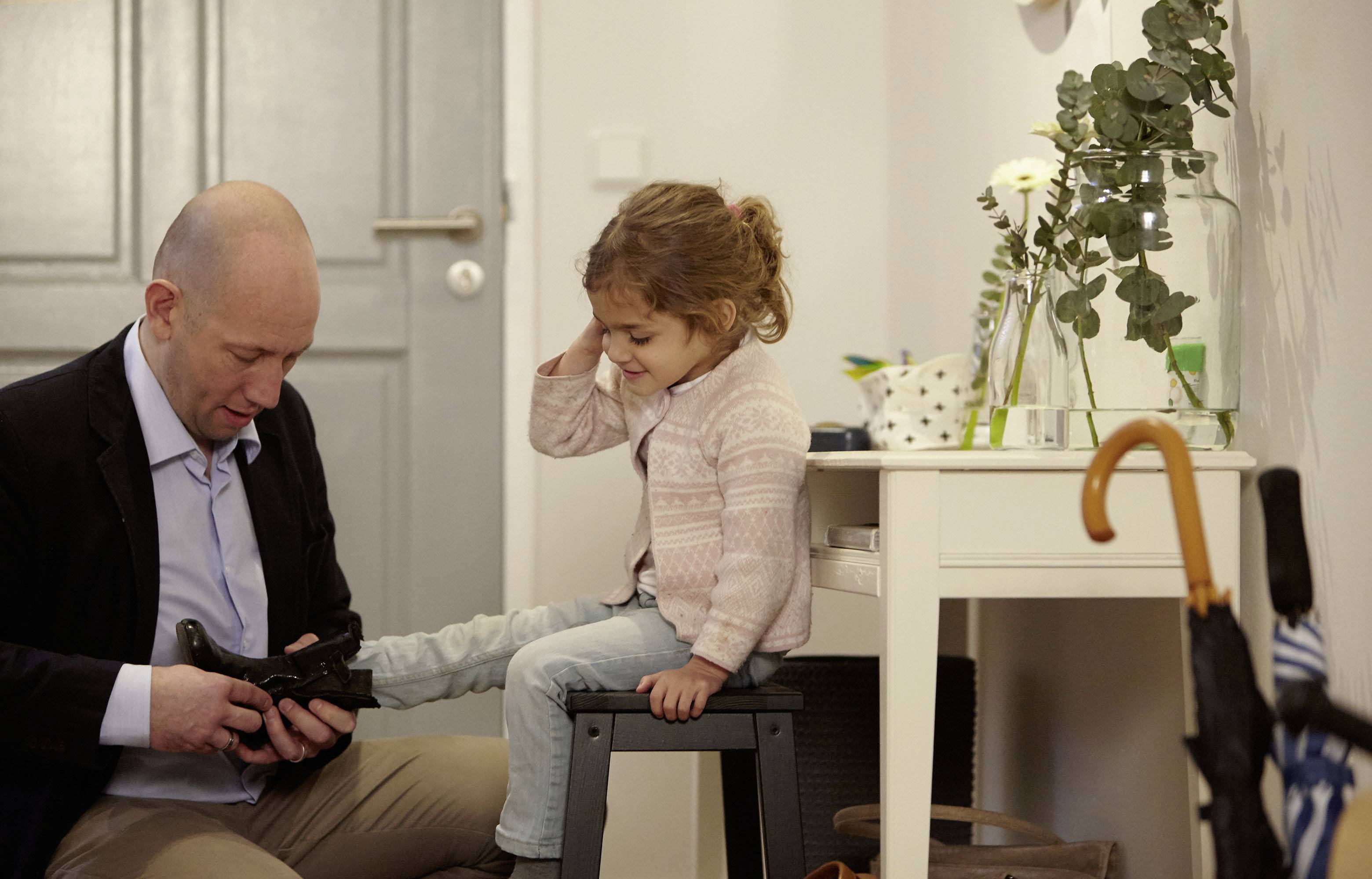 Un homme aide un enfant à mettre ses chaussures dans un couloir. À l'arrière-plan, on peut voir une table avec des fleurs et un parapluie.