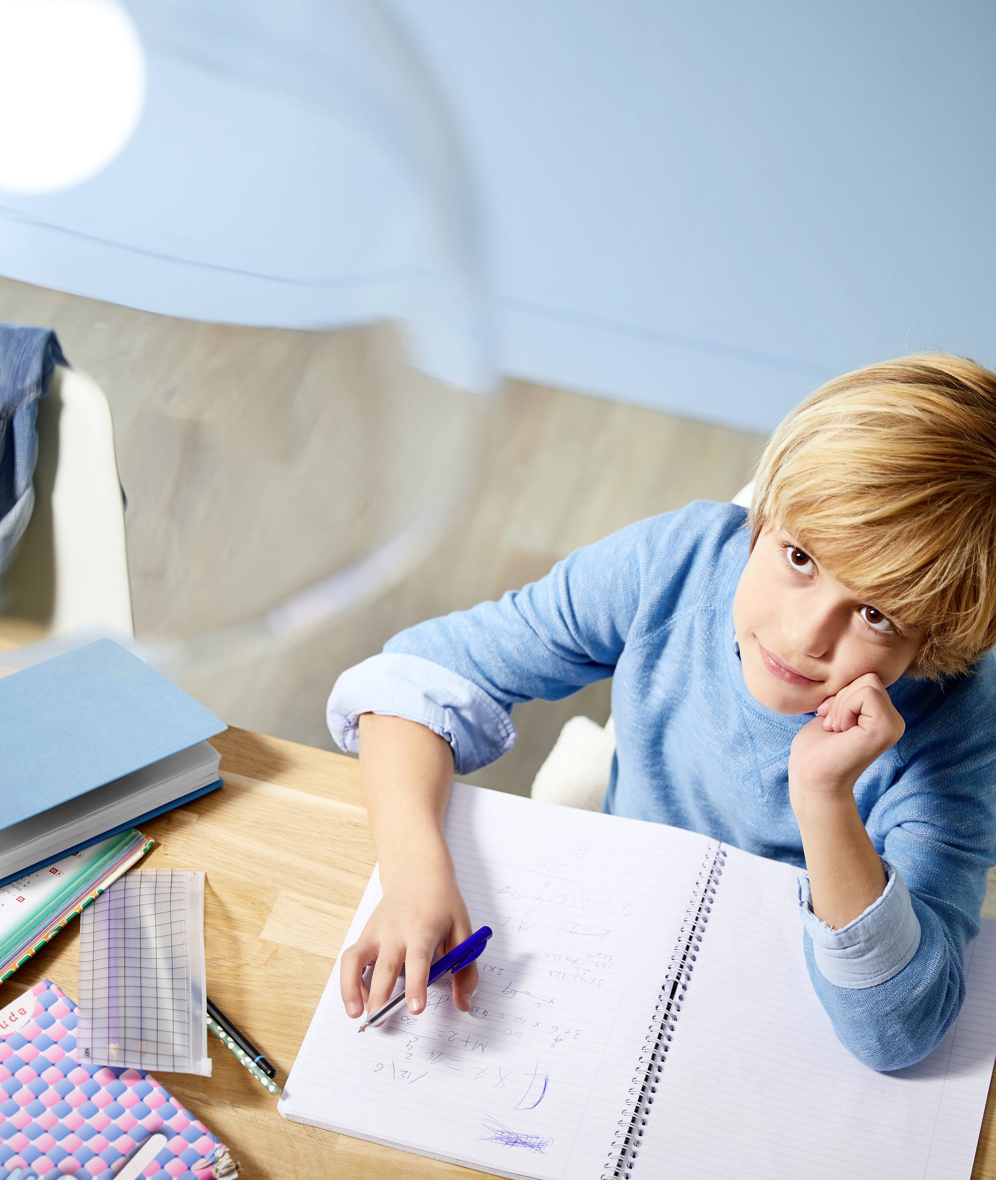Un garçon est assis à un bureau, écrit dans un cahier et regarde d'un air pensif. Des livres et des cahiers sont éparpillés en arrière-plan.
