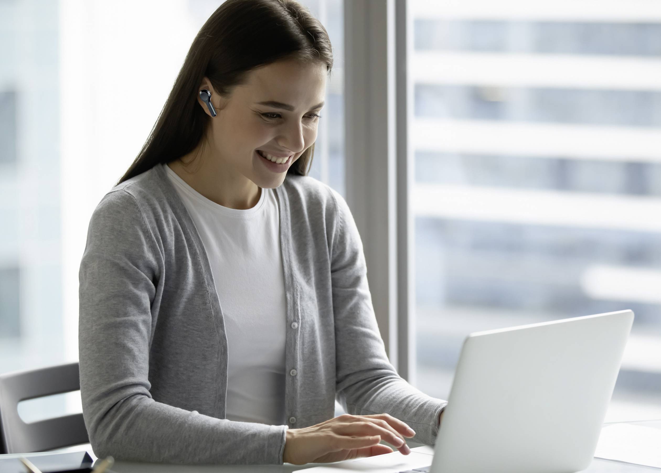 Une femme souriante, portant un casque, est assise devant un ordinateur portable dans un bureau moderne, absorbée par son travail.