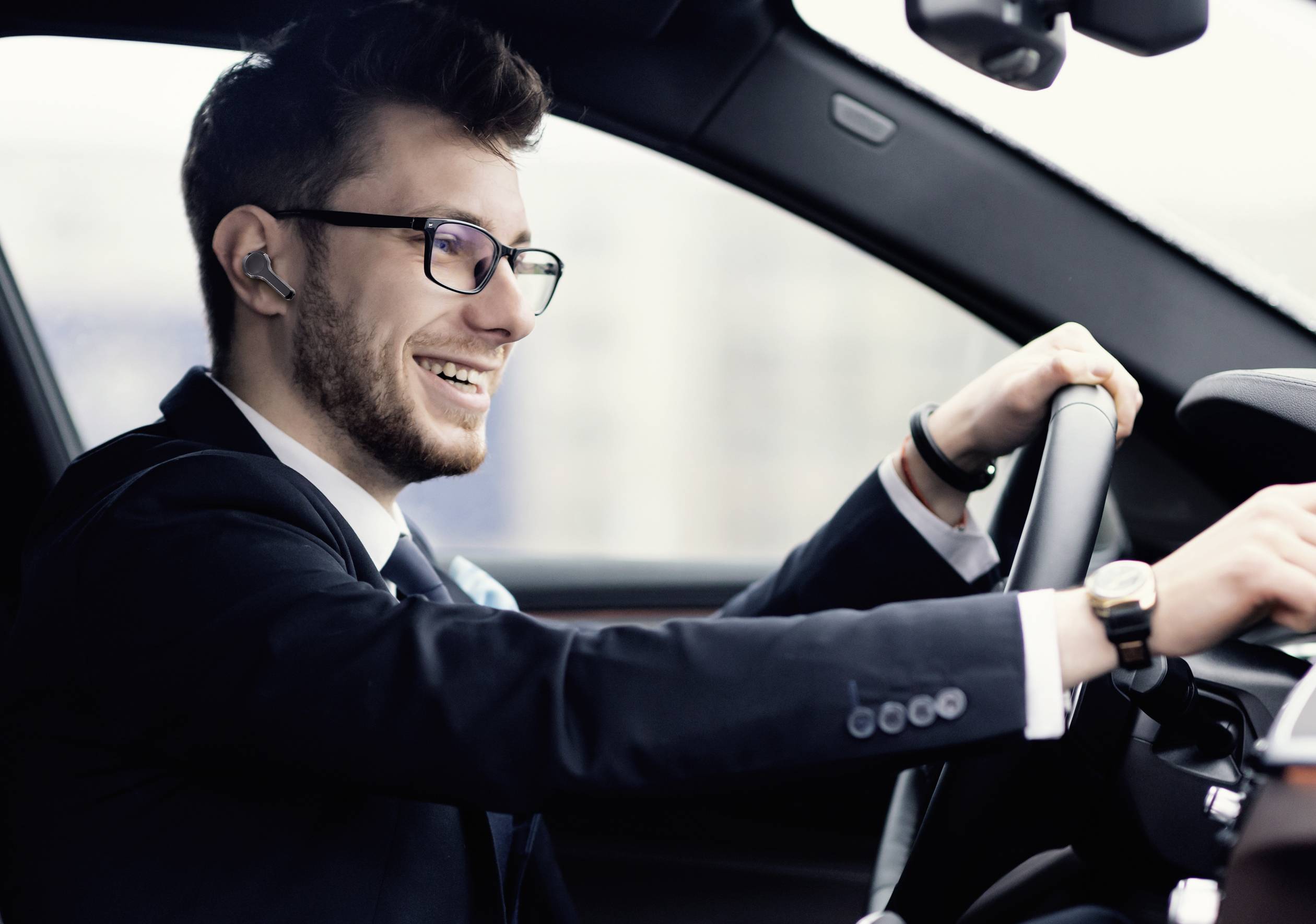 Un homme en costume et lunettes est assis souriant derrière le volant d'une voiture. Il semble satisfait ou ravi.