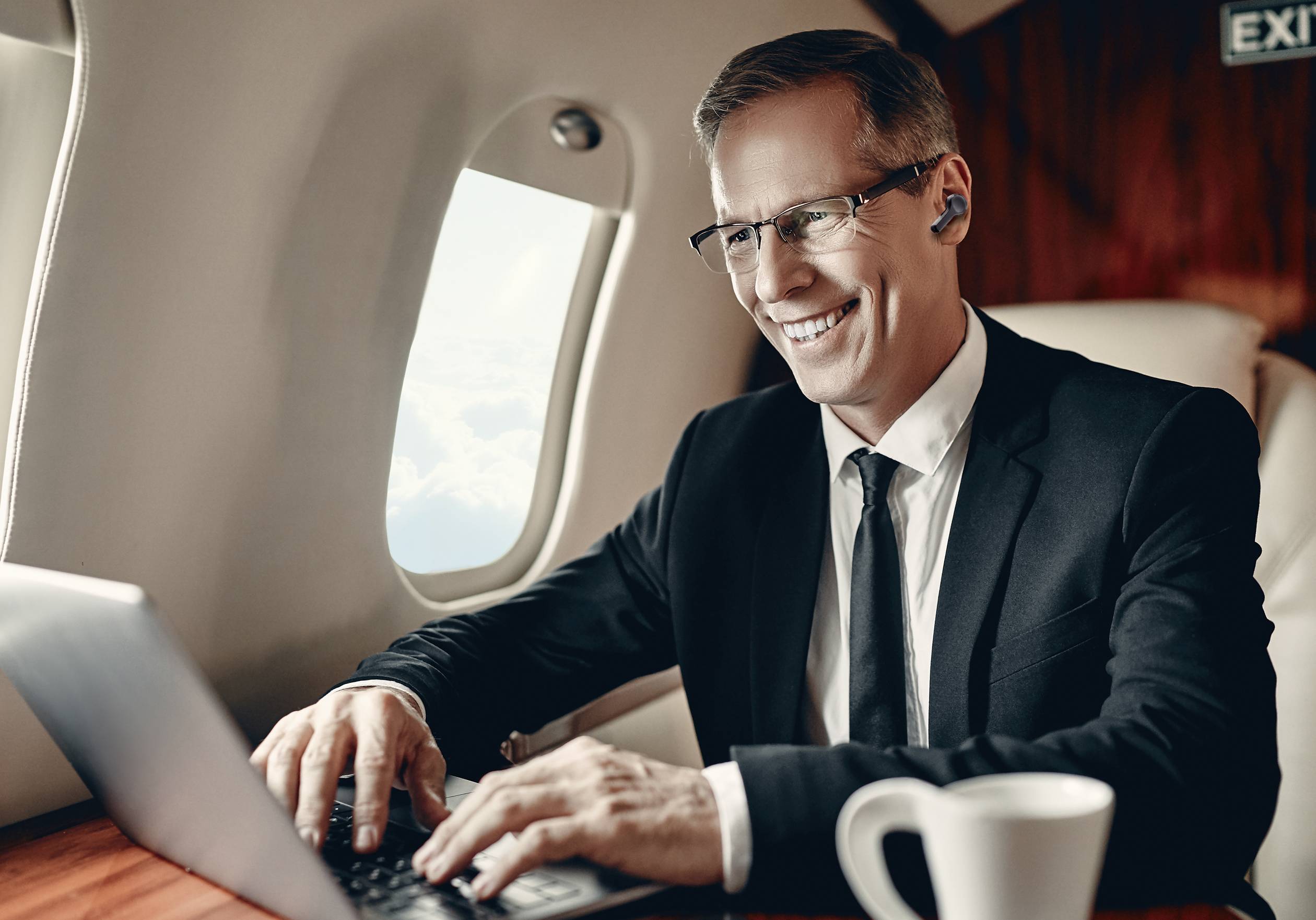 Un homme en costume travaille en souriant sur un ordinateur portable à l'intérieur d'un avion. Une tasse de café est posée sur la table à côté de lui.