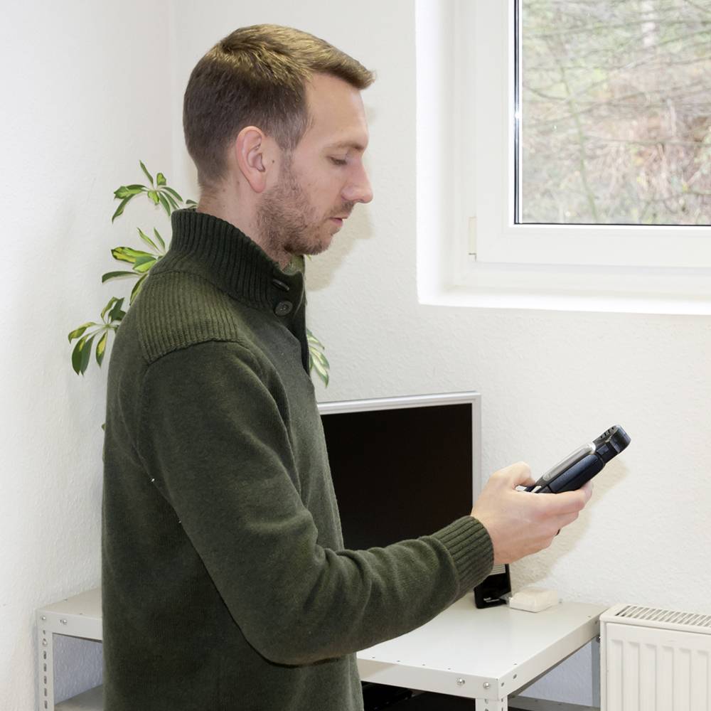 Un homme se tient dans une pièce, tenant un smartphone à la main et regardant son écran. À l'arrière-plan, on peut voir un moniteur et des plantes.