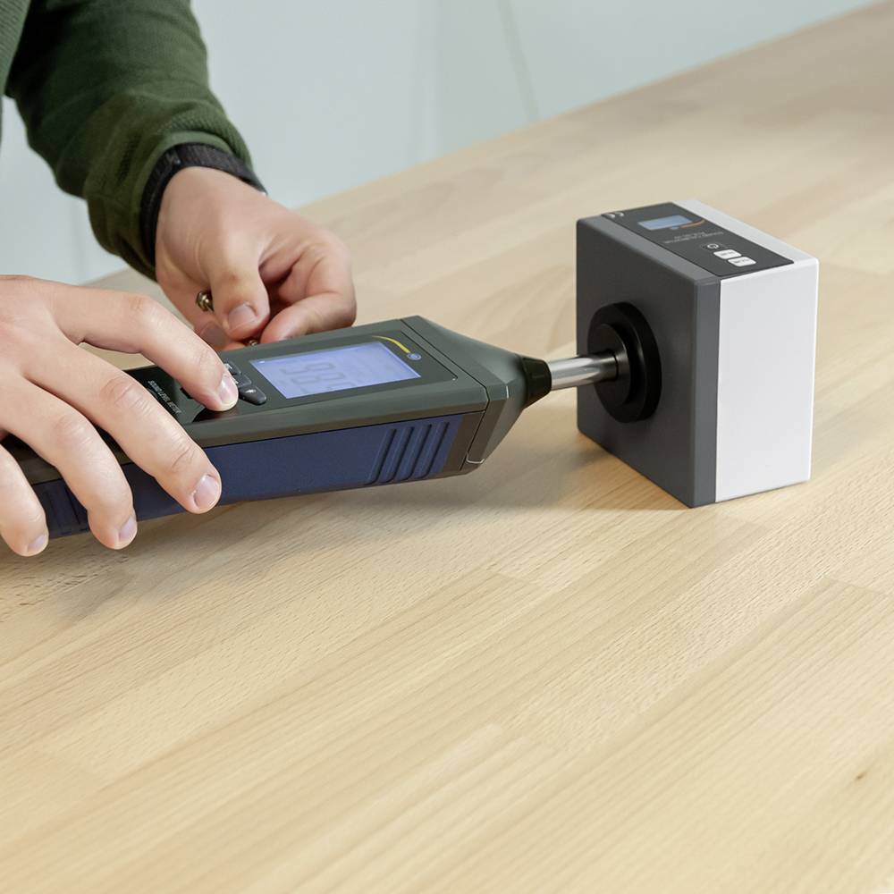 Une personne mesure la dureté d'un bloc de bois sur une surface de table à l'aide d'un appareil de mesure numérique.