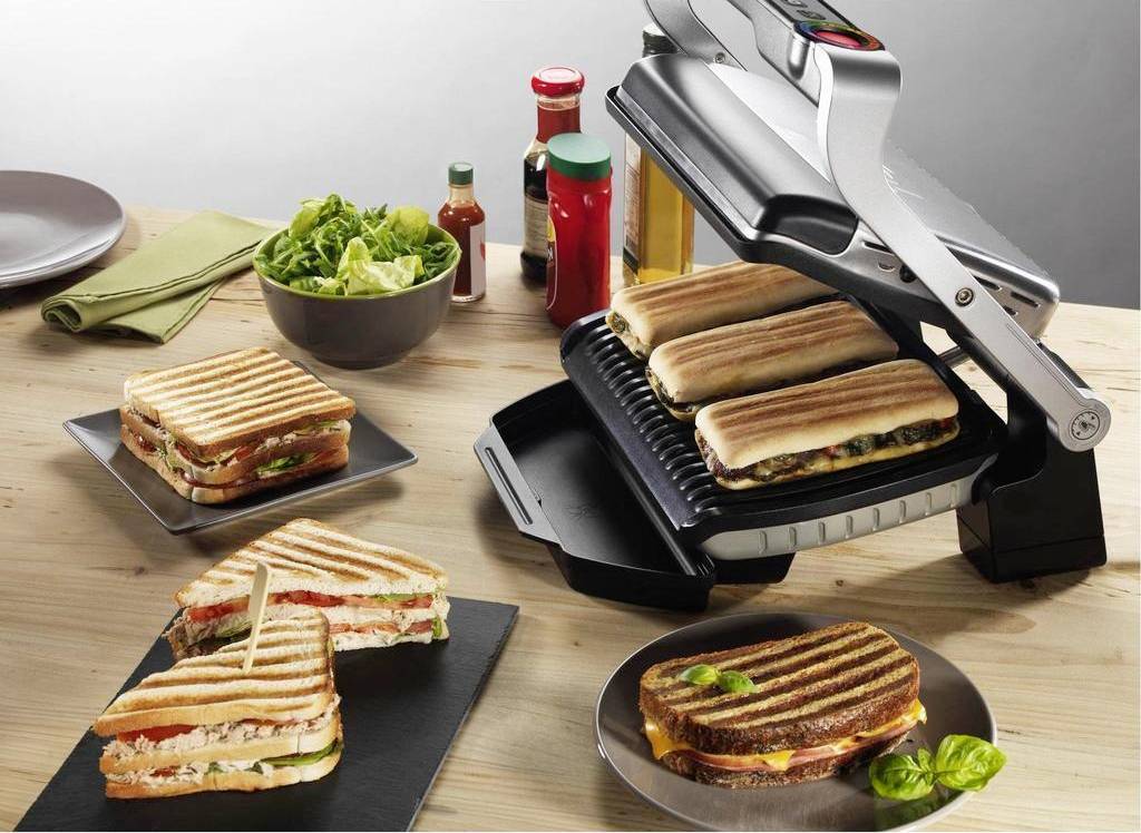 Une table avec un appareil de grillade qui fait griller plusieurs sandwichs. À côté, des assiettes contenant des sandwichs préparés et des bols de salade.