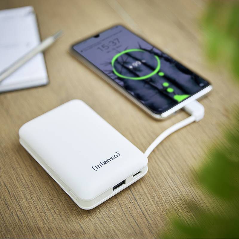 Une batterie externe charge un smartphone posé sur une table en bois. Un stylo et un carnet de notes sont également visibles.