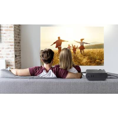 Un couple est assis sur un canapé et regarde un film projeté sur un écran, montrant une famille qui court joyeusement à travers un champ.