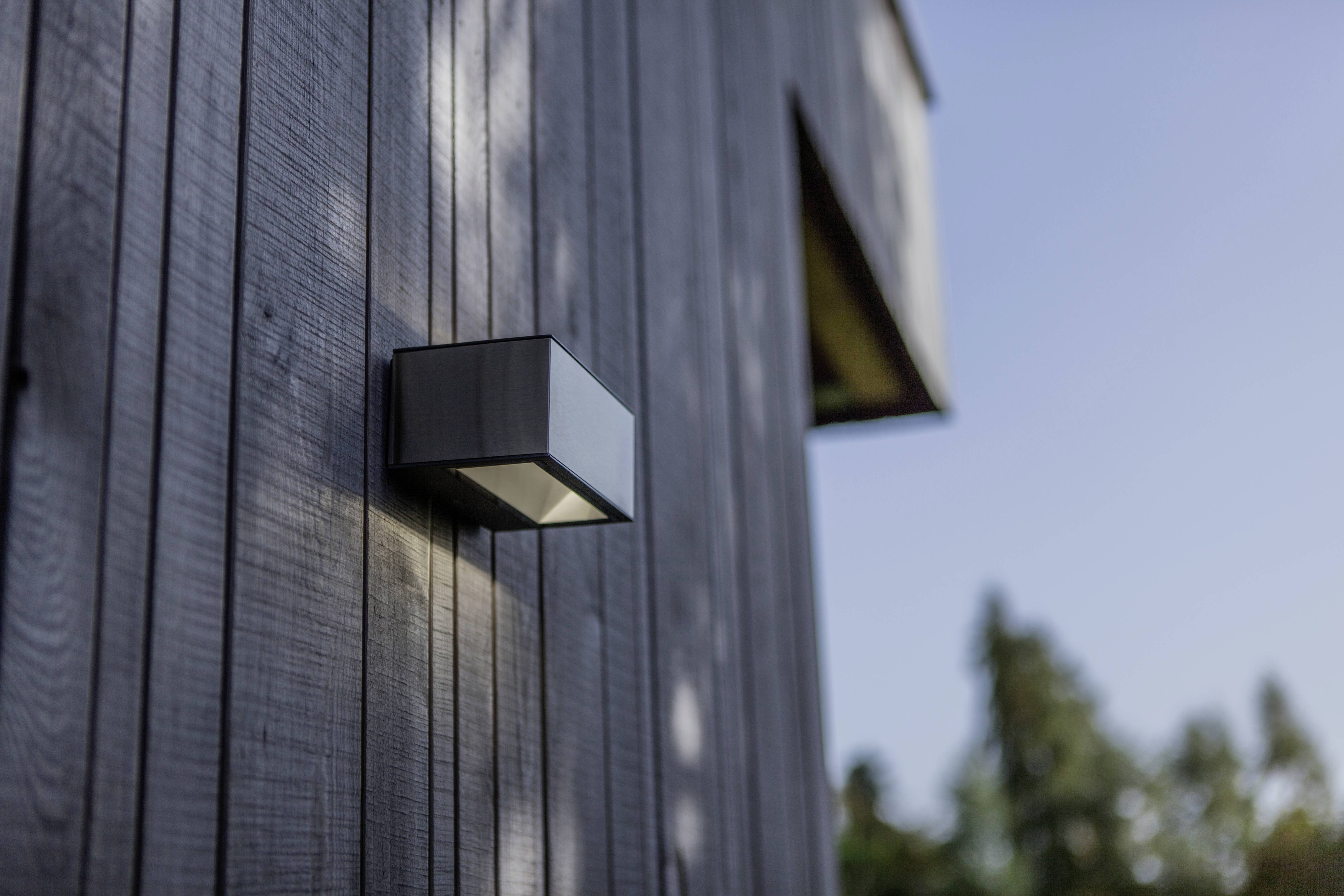 Luminaire extérieur fixé sur un mur en bois d'un bâtiment moderne, entouré d'arbres en arrière-plan, éclairé à la lumière du jour.