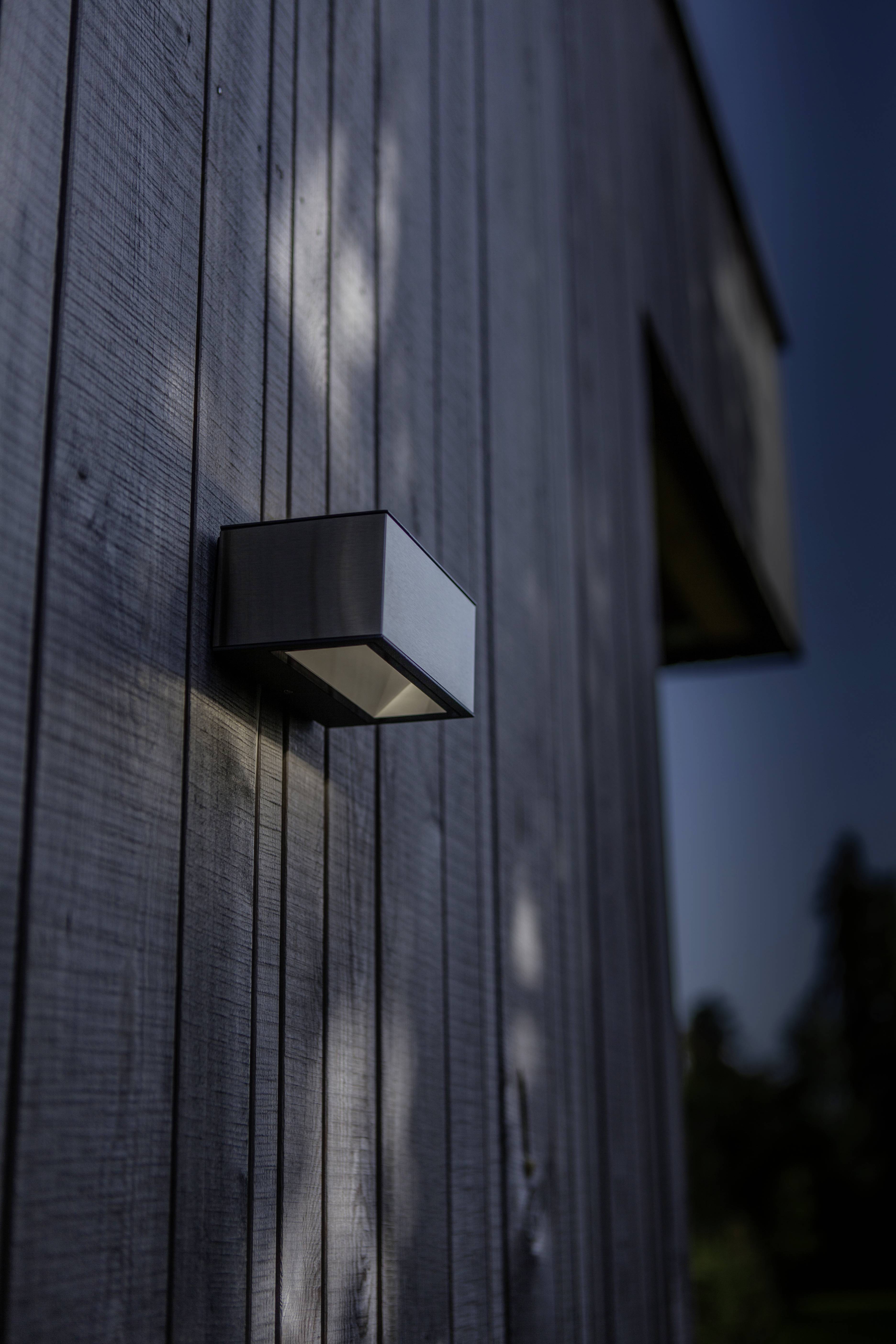 Mur extérieur d'une façade en bois avec un luminaire contemporain projetant une lumière douce sur la structure ; des arbres visibles en arrière-plan.
