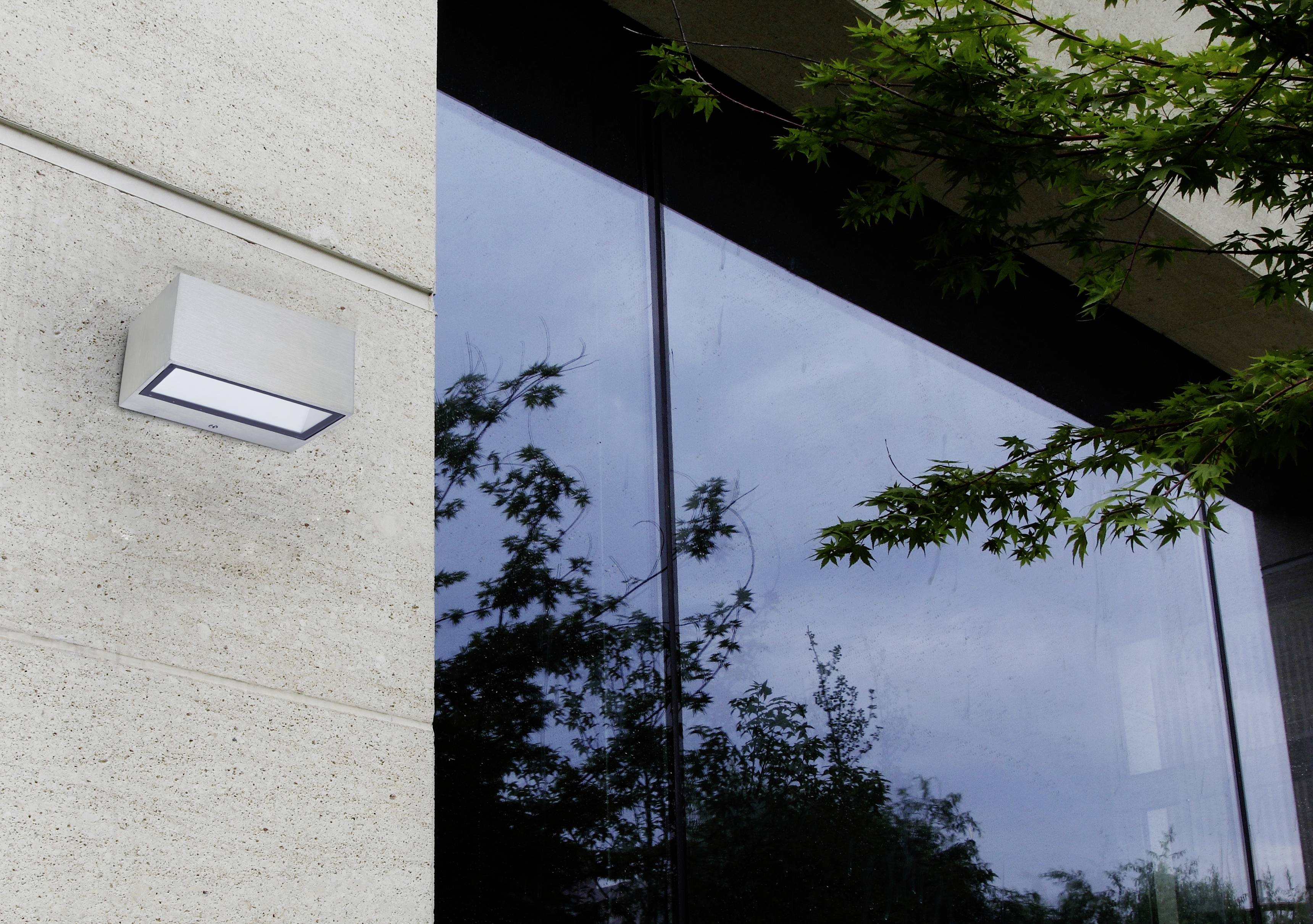 Vue extérieure d'un bâtiment avec façade vitrée et applique murale moderne. Des branches d'arbres se reflètent dans la fenêtre.