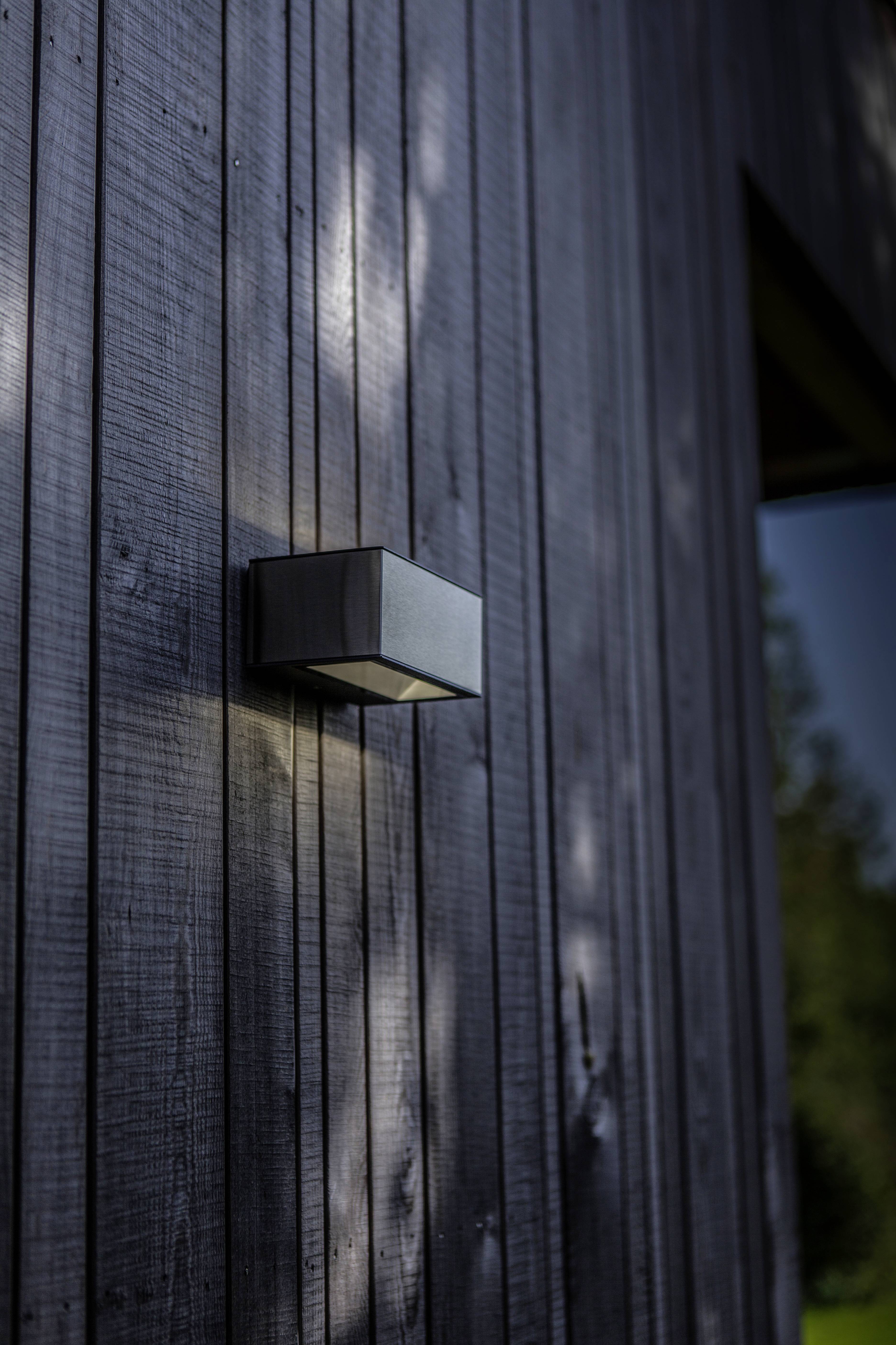 Une lampe extérieure moderne sur une façade en bois sombre d'un bâtiment, éclairée à la lumière du jour. Des arbres sont visibles en arrière-plan.