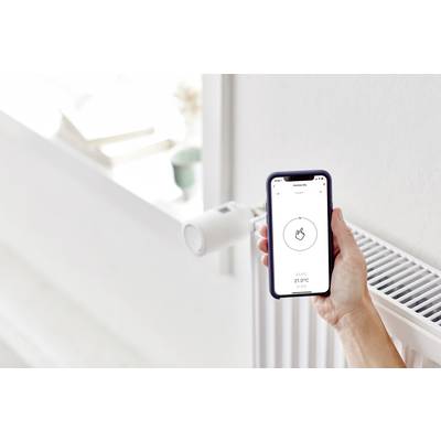 Une main tient un smartphone avec une application de contrôle du chauffage. En arrière-plan, on aperçoit un radiateur équipé d'un thermostat intelligent.