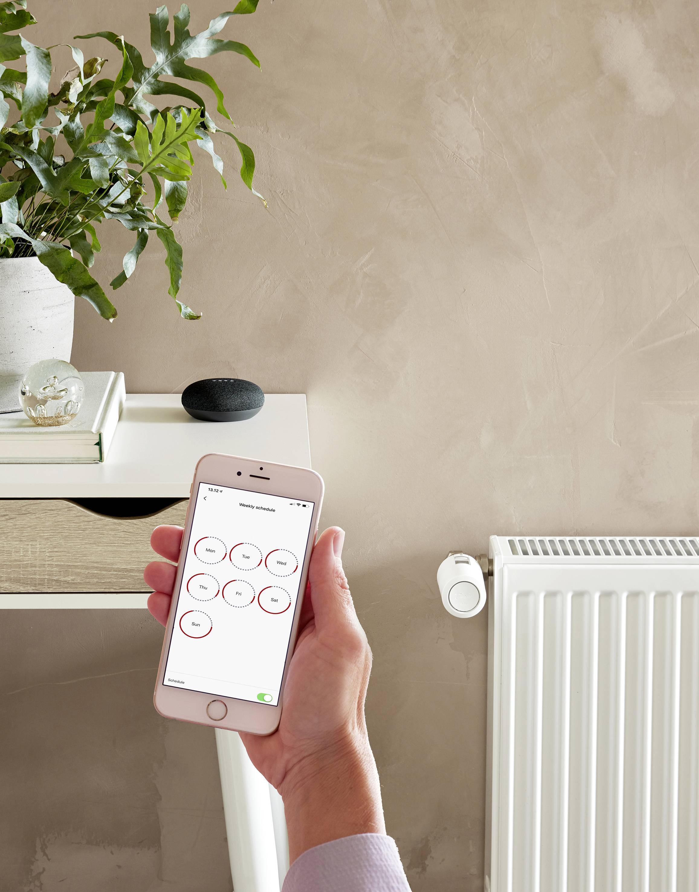 Une personne contrôle la température des radiateurs avec un smartphone dans une pièce moderne. Une table et une plante sont en arrière-plan.