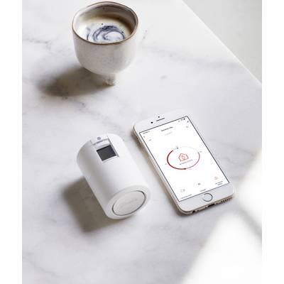 Une tasse de café blanche, un smartphone avec une application ouverte et un appareil cylindrique blanc sont posés sur une table en marbre.