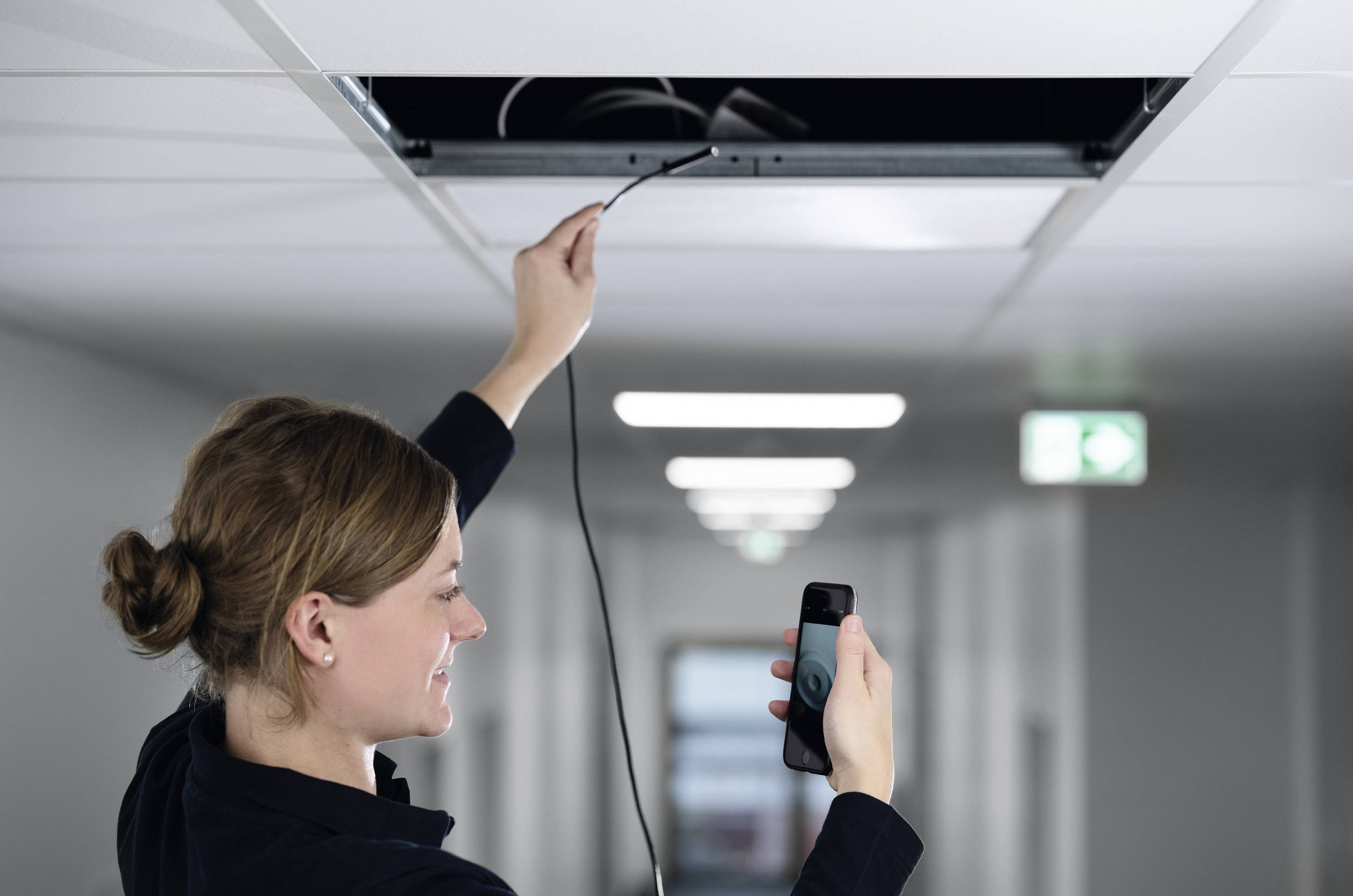Une personne se tient sur une échelle et vérifie des câbles suspendus à partir d'une ouverture au plafond à l'aide d'un smartphone.