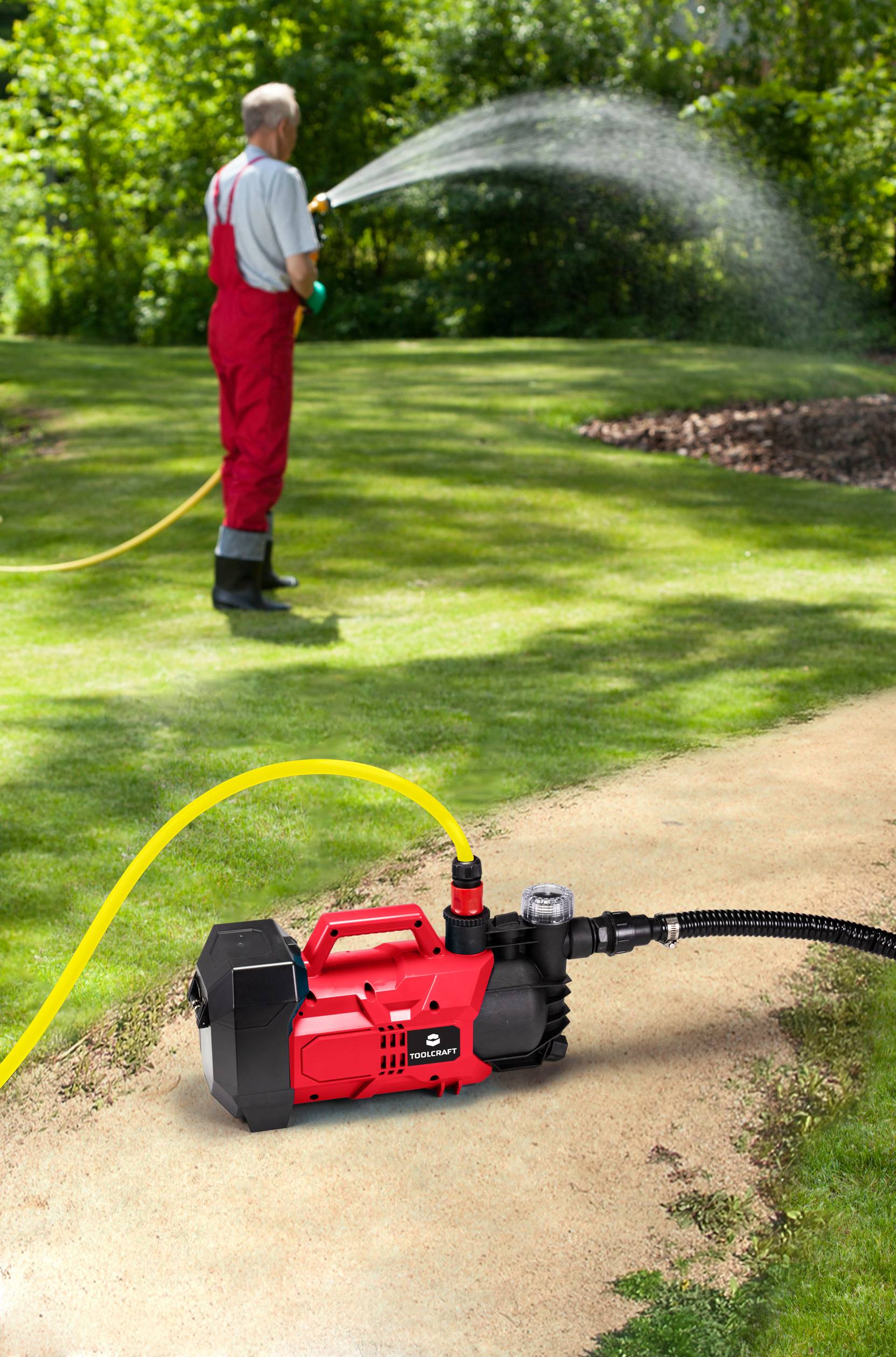 Un jardinier en tenue rouge arrose la pelouse avec un tuyau. Au premier plan, une pompe à eau rouge est posée sur un chemin.