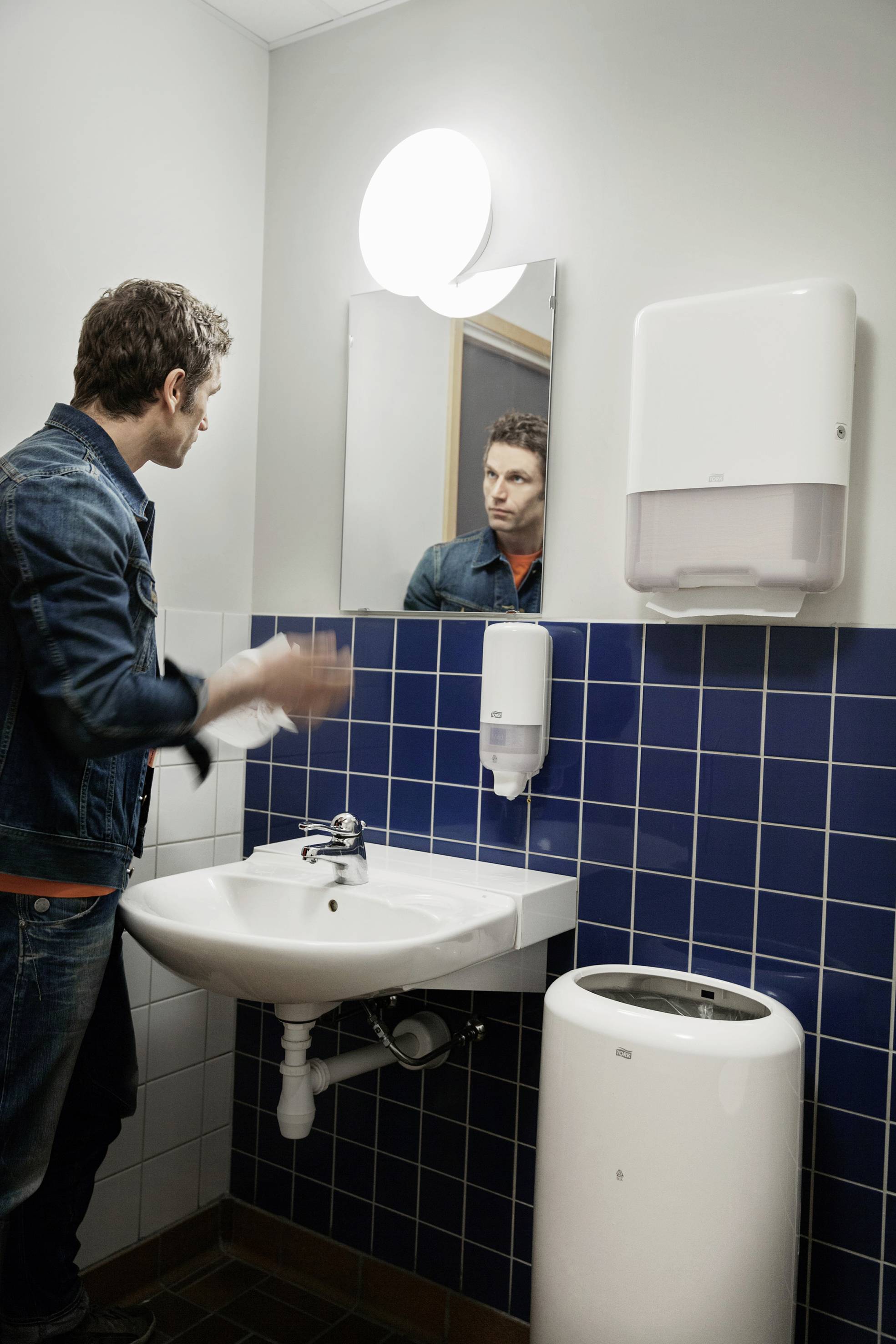 Un homme se lave les mains dans une salle de bain carrelée de bleu. Un miroir, un distributeur de savon et un distributeur d'essuie-tout sont visibles.