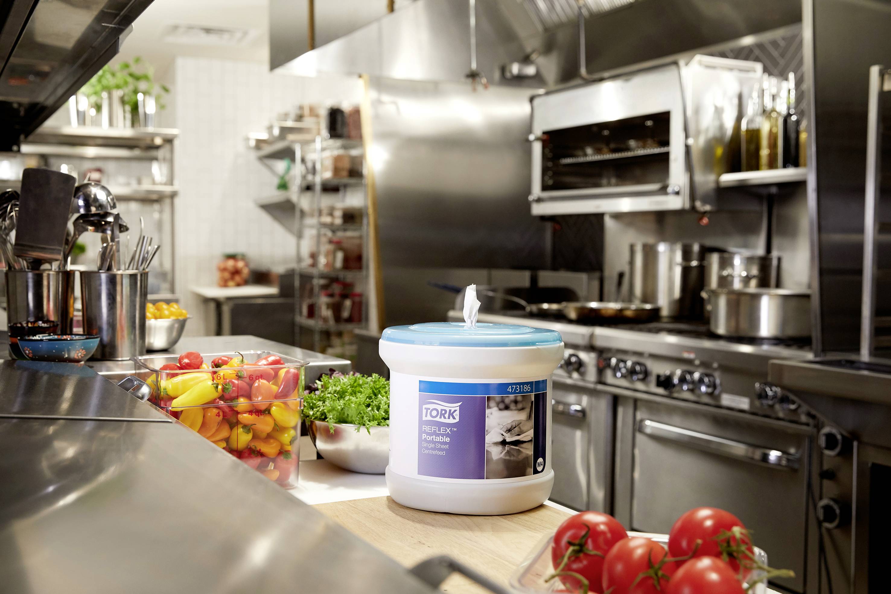 Une cuisine professionnelle de restaurant avec des appareils en acier inoxydable. Au premier plan se trouvent un seau d'essuie-tout, des bandelettes de poivrons colorées et des tomates.