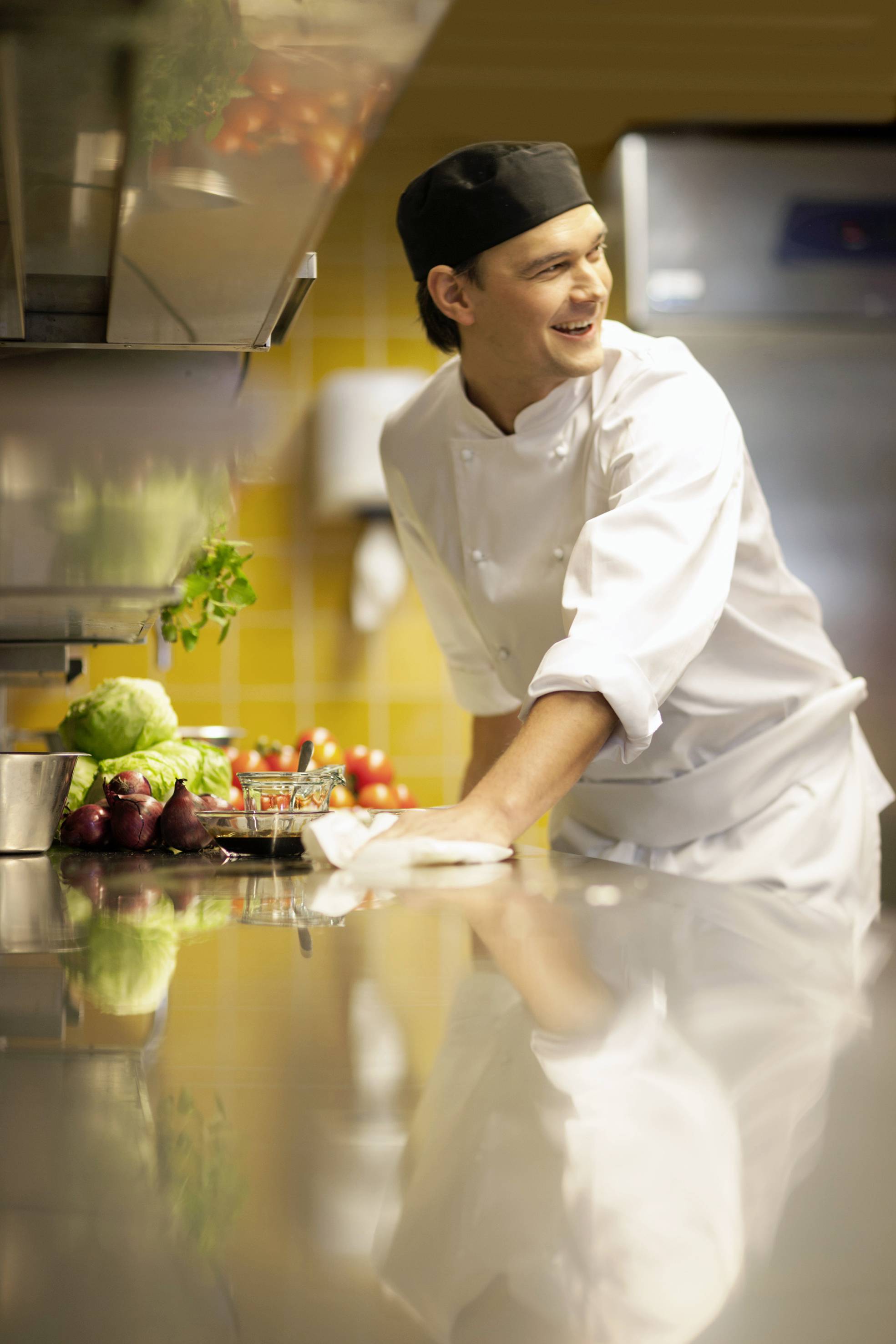 Un chef dans son uniforme blanc et sa toque noire sourit dans une cuisine professionnelle, entouré de légumes frais.