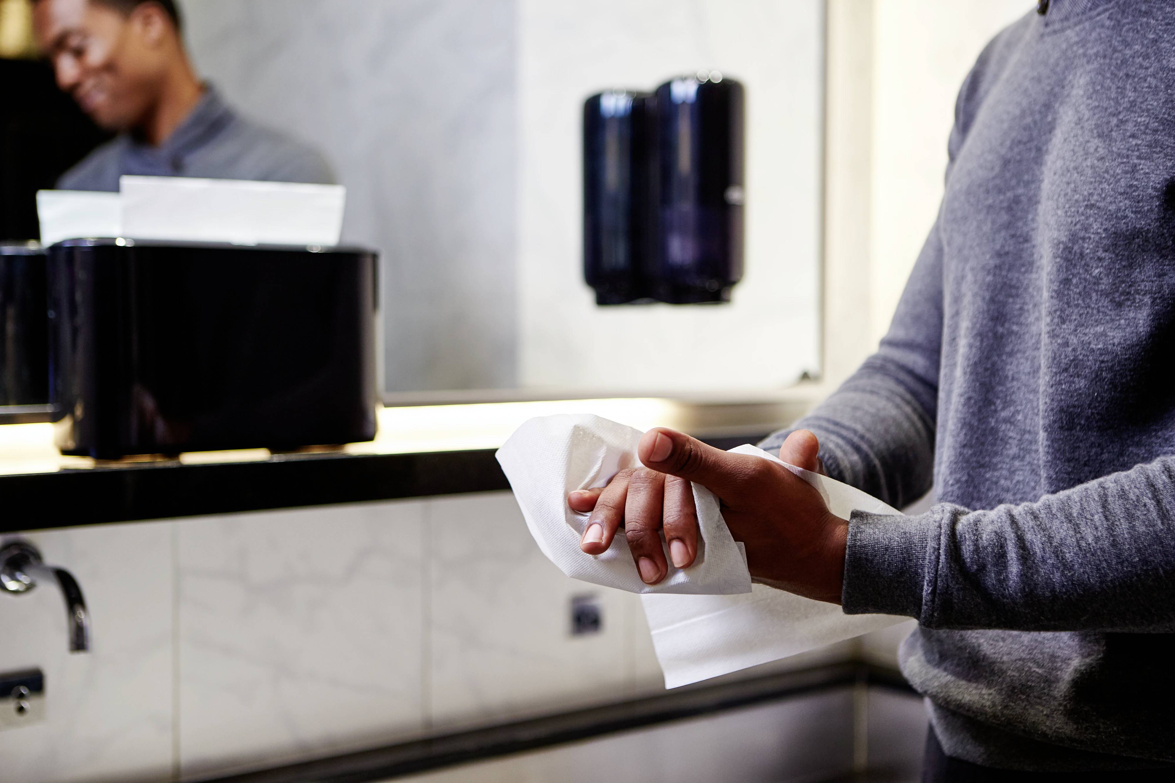 Une personne s'essuie les mains avec un essuie-papier dans une salle de bain. Un miroir et un distributeur de savon ou d'essuie-mains sont visibles en arrière-plan.