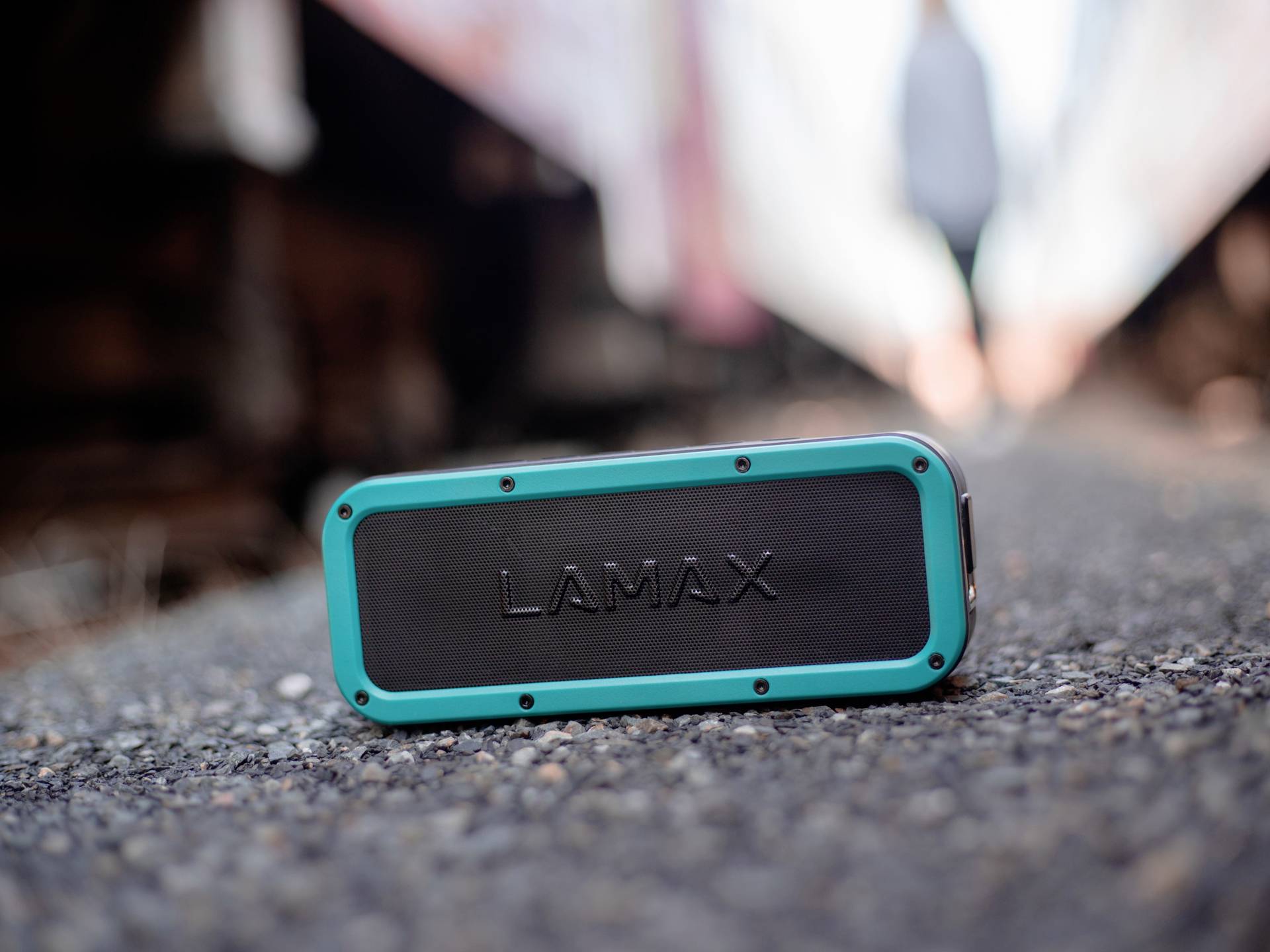 Enceinte portable Lamax sur une surface asphaltée en extérieur. L'arrière-plan flou suggère un environnement extérieur.