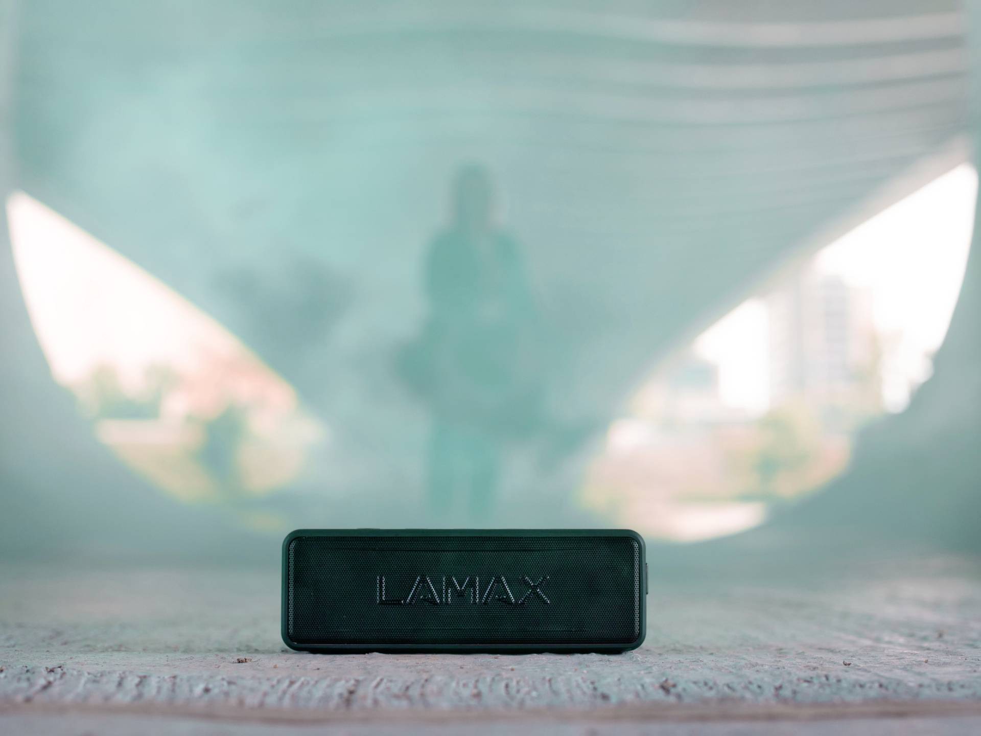 Un haut-parleur noir rectangulaire portant l'inscription 'LAMAX' est posé sur une surface en béton dans un tunnel urbain. Une silhouette de personne est visible en arrière-plan.