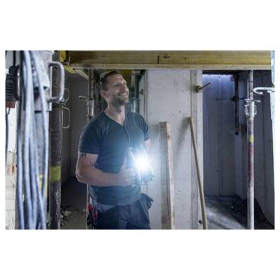 Travailleur souriant vêtu d'une chemise vert foncé tenant une lampe de poche brillante dans une zone de construction inachevée.
