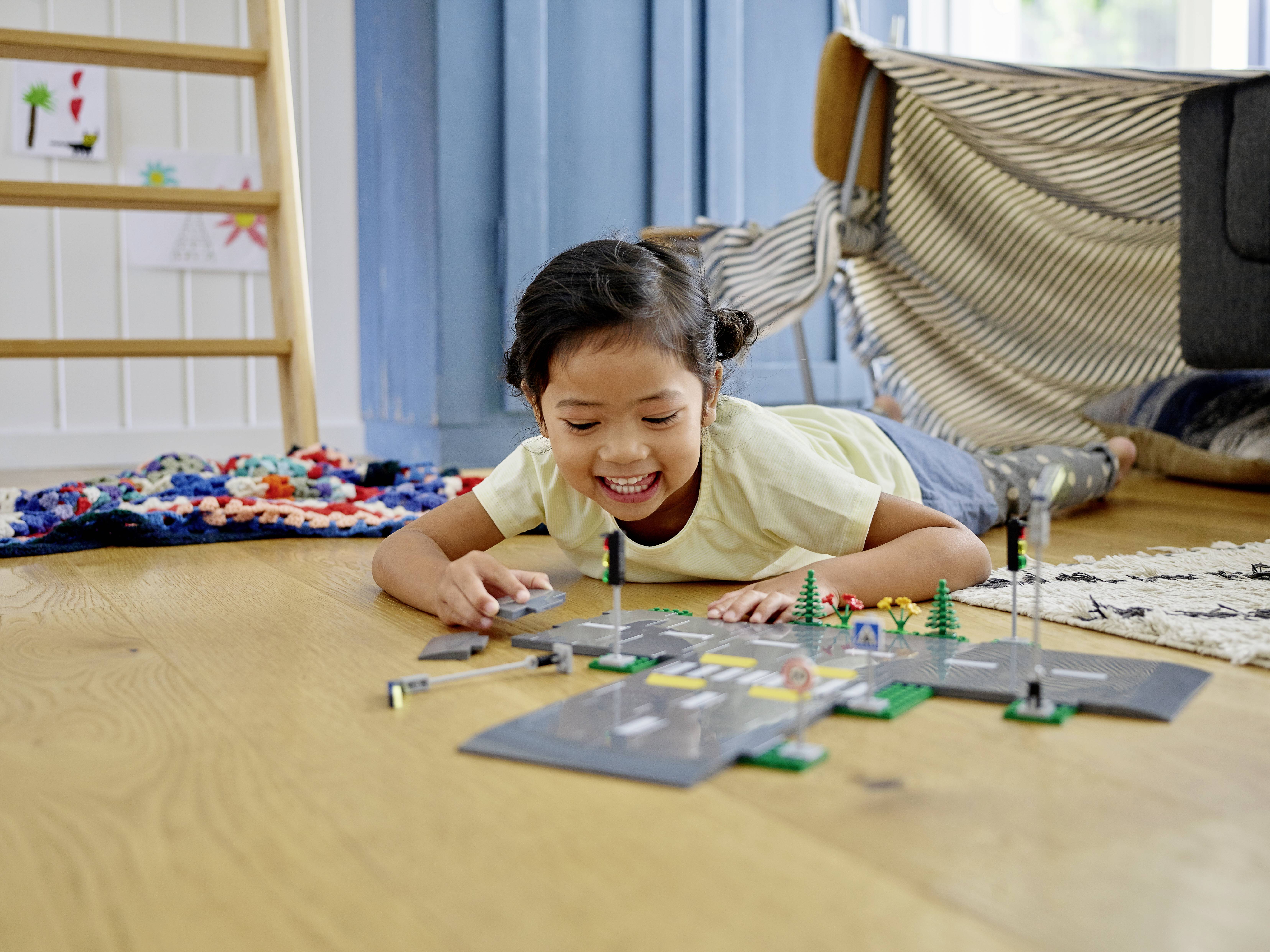 Un enfant joue en souriant avec un jouet Lego sur le plancher en bois du salon. En arrière-plan, on aperçoit un tapis et un meuble transformé en tente.