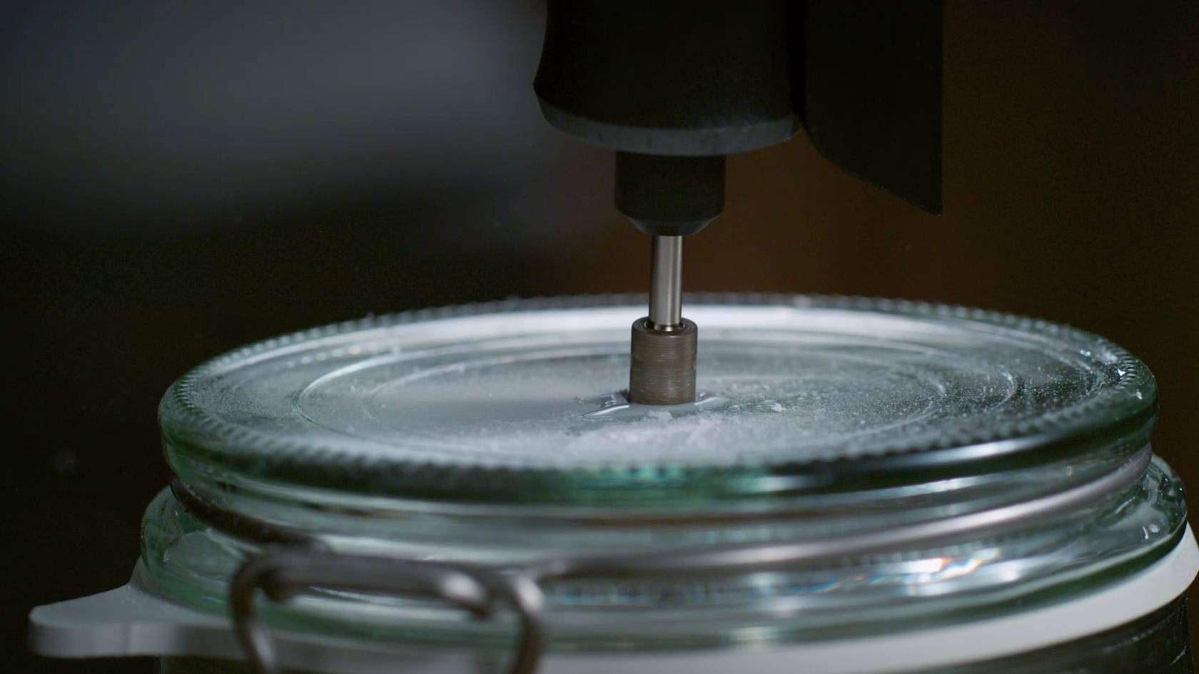 Un foret métallique perce un trou dans un couvercle en verre.