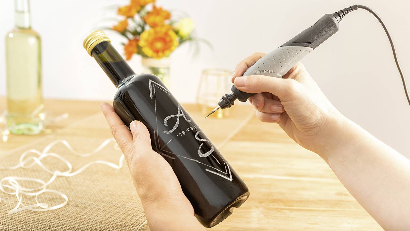 Une personne grave un motif sur une bouteille de verre sombre à l'aide d'un outil. À l'arrière-plan, du vin est posé sur une table décorée de fleurs.