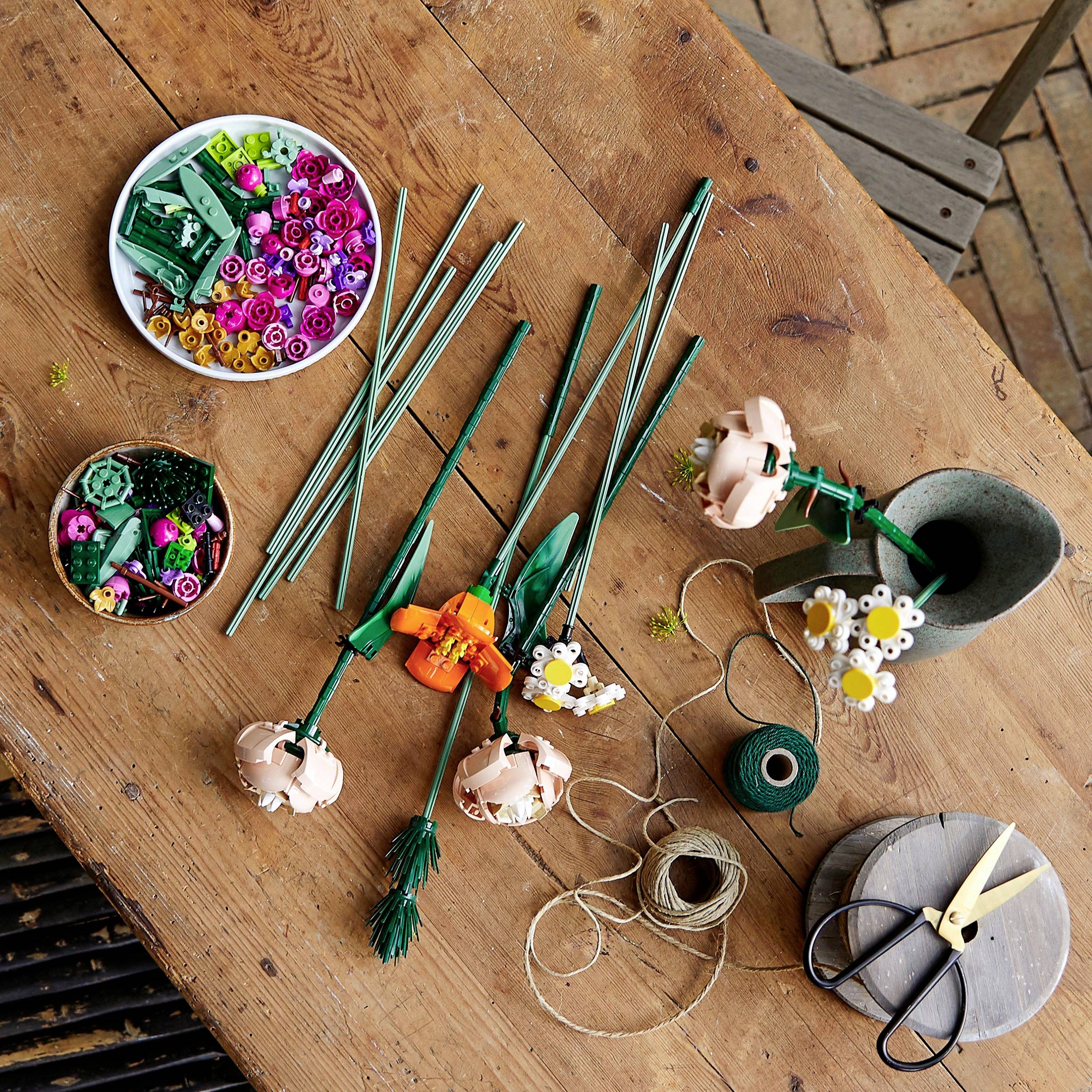 Table de bricolage avec différentes fleurs en blocs de construction, tiges, pétales, ciseaux, fil et arrosoir sur surface en bois.