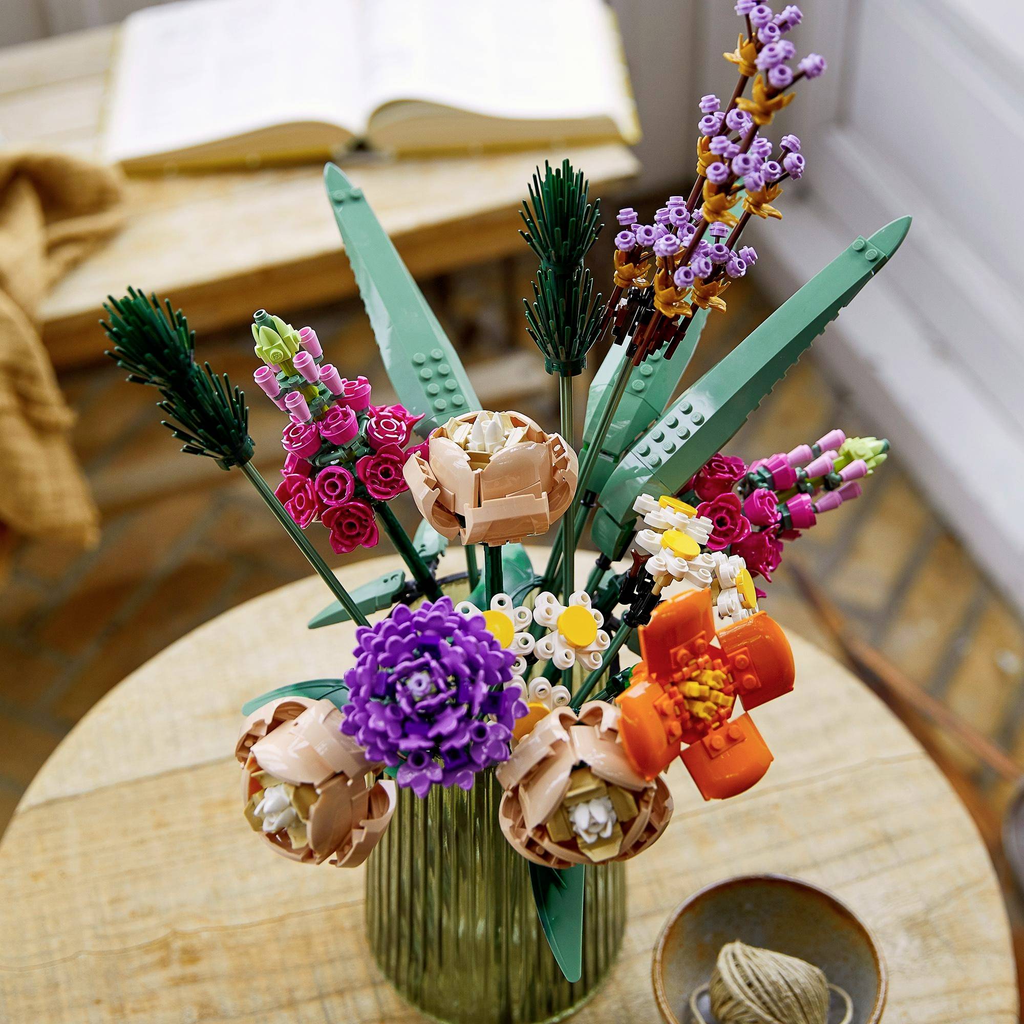 Un bouquet de fleurs LEGO colorées est placé dans un vase sur une table en bois. À l'arrière-plan, on peut voir un livre et un tissu.