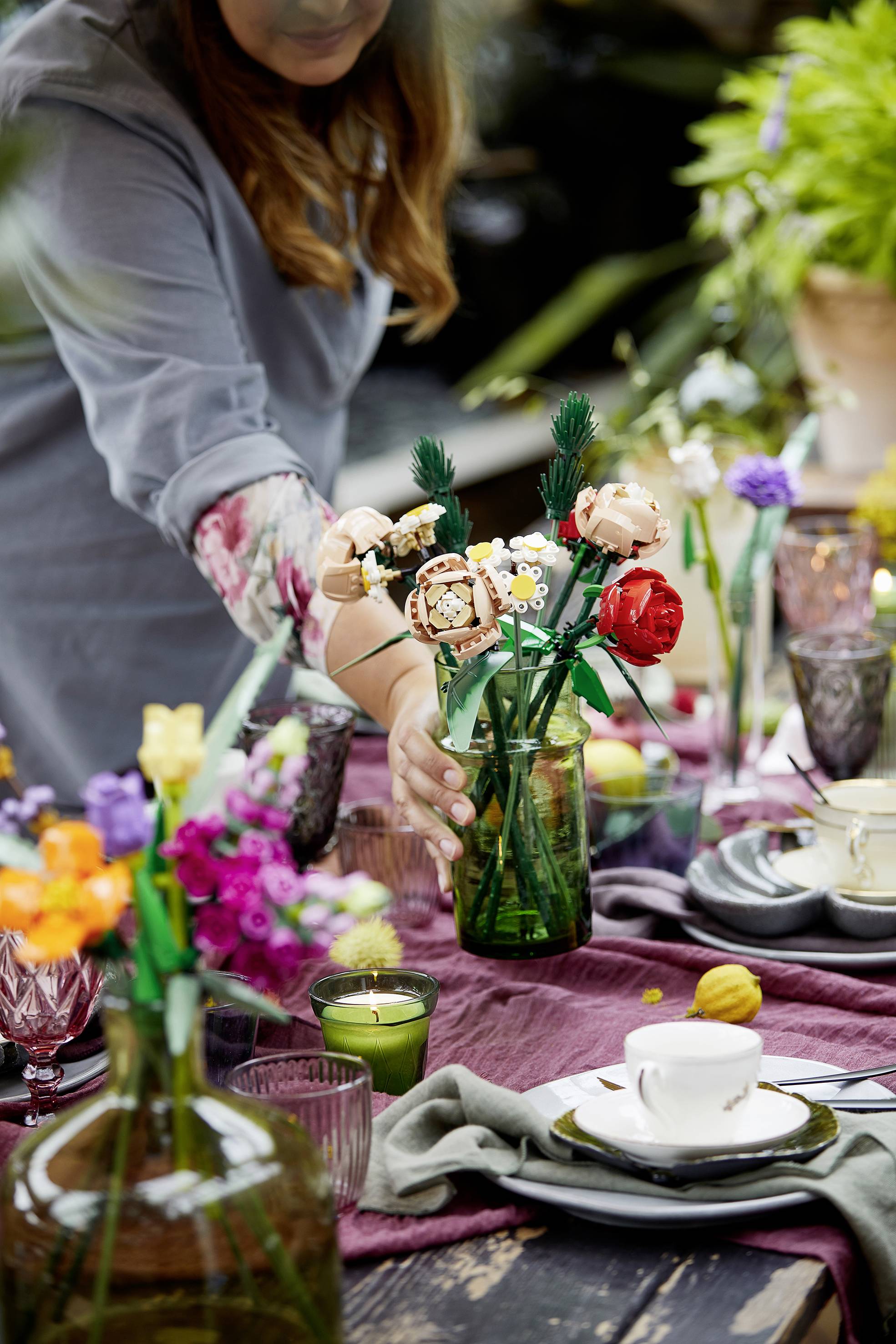 Une personne dispose un bouquet de fleurs dans un vase sur une table décorativement dressée avec des tasses, des assiettes et des bougies.