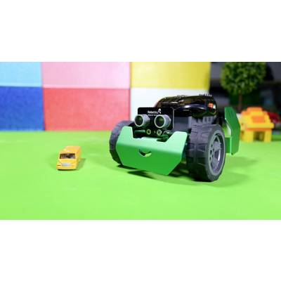 Robot jouet au visage joyeux roulant sur une surface verte à côté d'une petite voiture jouet jaune. Mur coloré en arrière-plan.