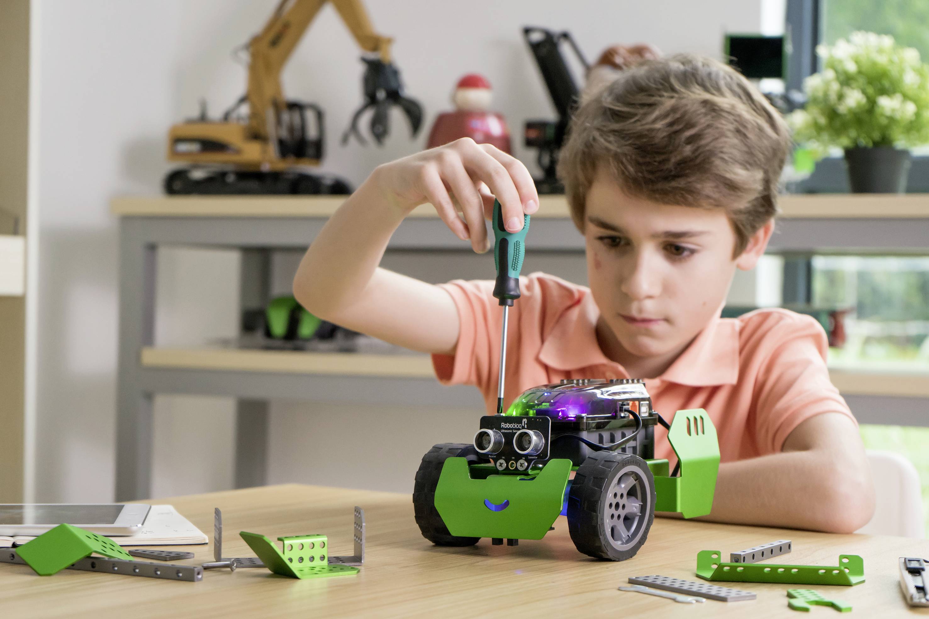 Un enfant portant une chemise orange assemble une voiture jouet avec un tournevis. Des jouets sont visibles en arrière-plan.