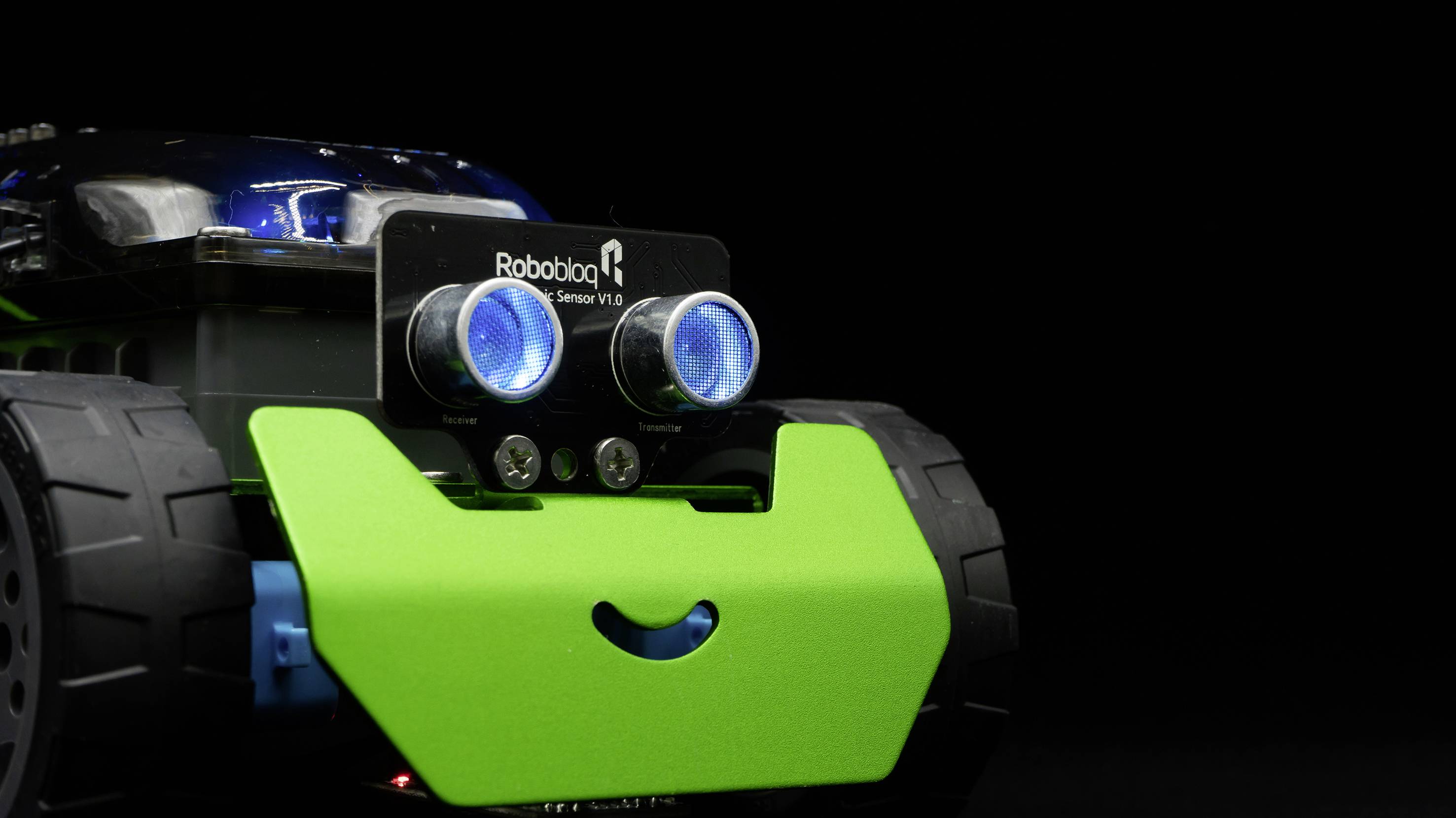 Un petit véhicule de jouet, ressemblant à un robot, avec de grands capteurs bleus à l'avant et un bouclier frontal vert souriant, est au centre de l'attention.