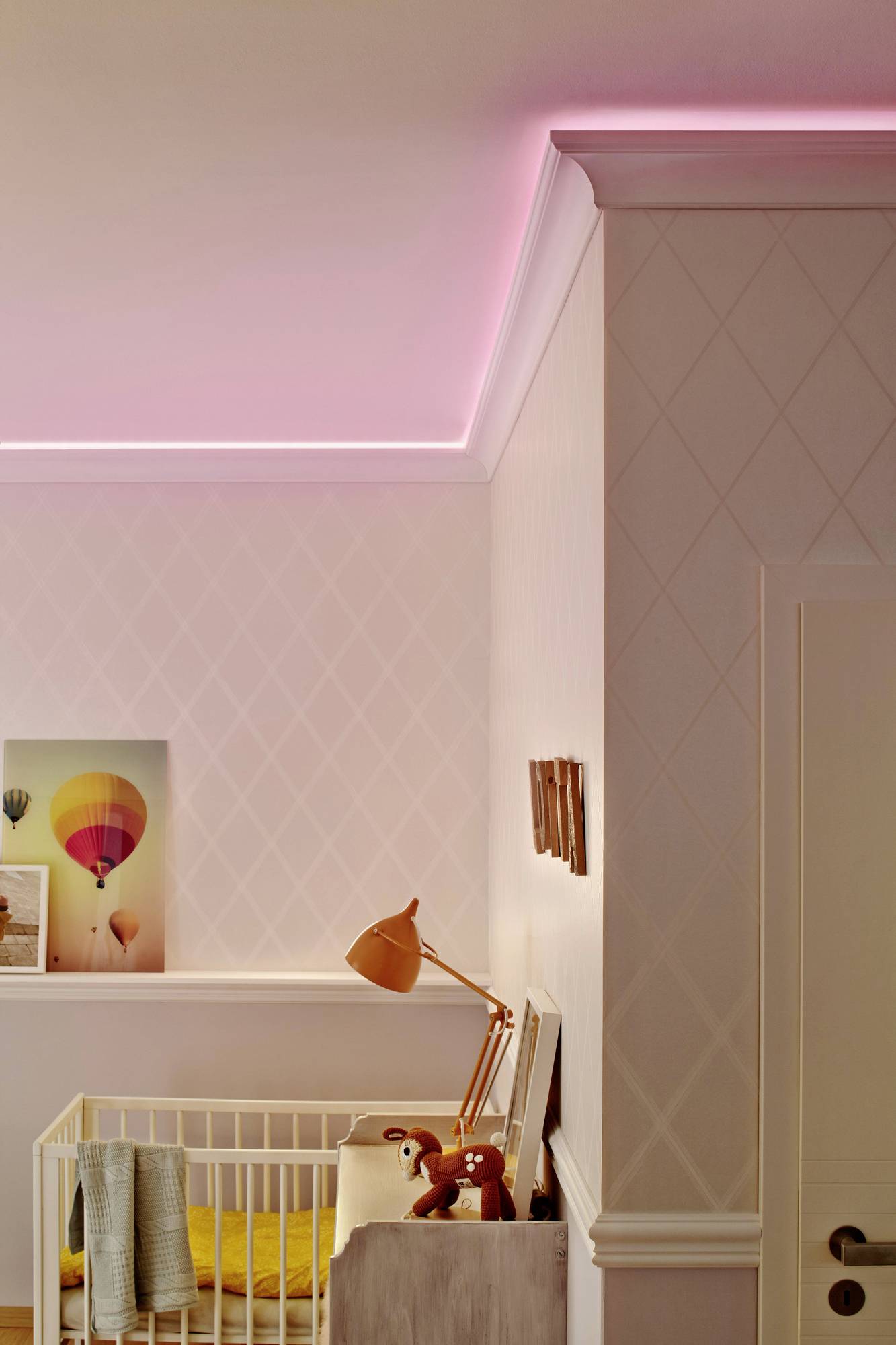 Chambre d'enfant avec un lit à barreaux blanc, de la literie jaune, des jouets et des œuvres d'art murales. Un éclairage LED rose illumine le plafond.