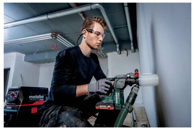 Une personne portant des lunettes de protection et des gants utilise une perceuse Metabo pour travailler sur un mur dans un environnement de construction.