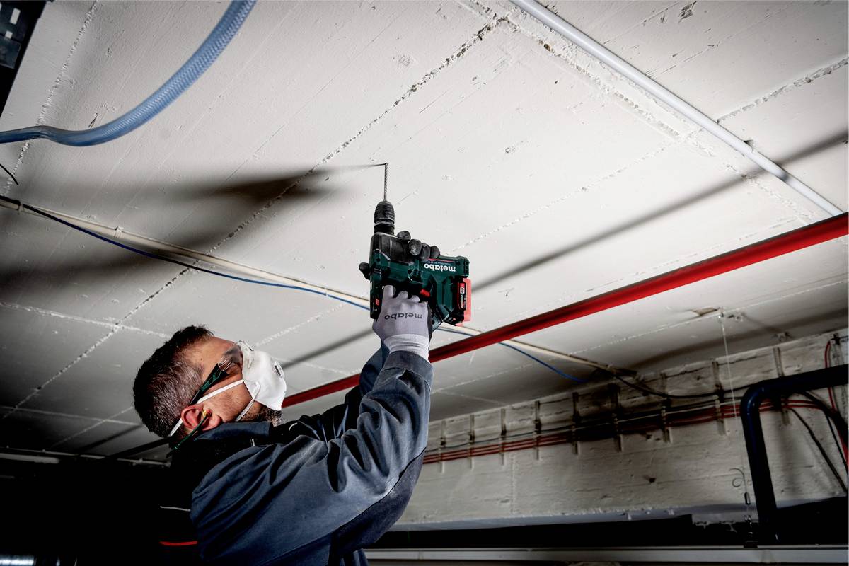 Une personne fore un plafond en béton à l'aide d'une perceuse. Elle porte des lunettes de protection et un masque respiratoire. Des câbles sont disposés le long du plafond.