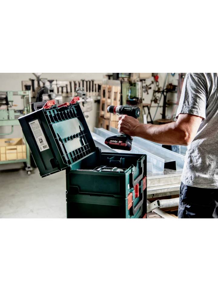 Une personne utilise une perceuse sans fil à côté d'une boîte à outils ouverte dans un atelier. Des machines et des étagères sont visibles en arrière-plan.