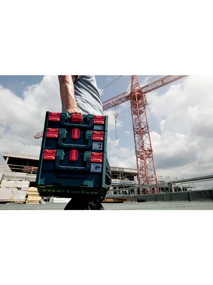Une personne transporte une valise à outils Metabo sur un chantier de construction, avec une grue et un bâtiment en cours de construction visibles en arrière-plan.