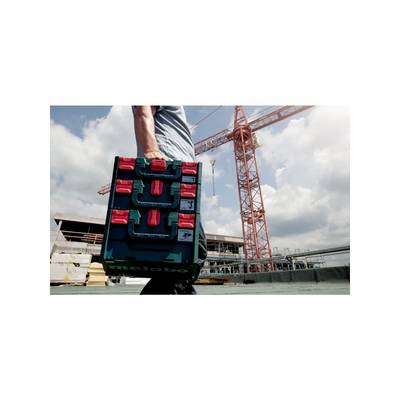 Une personne transporte une valise à outils Metabo sur un chantier de construction, avec une grue et un bâtiment en cours de construction visibles en arrière-plan.