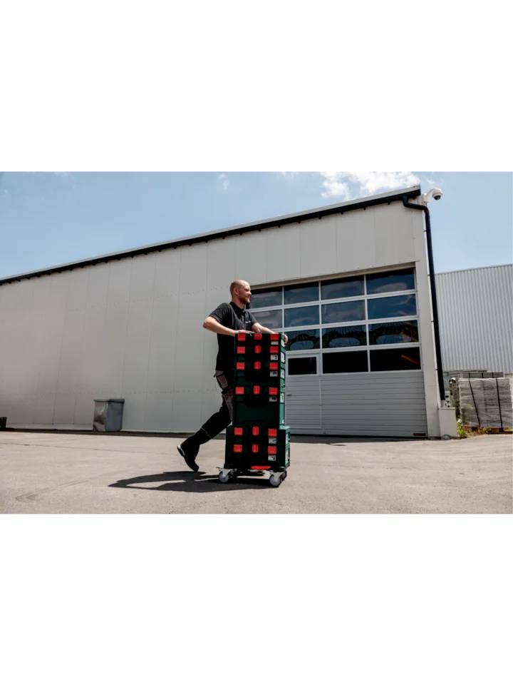 Un homme tire un chariot chargé de caisses empilées devant un grand entrepôt par temps ensoleillé.