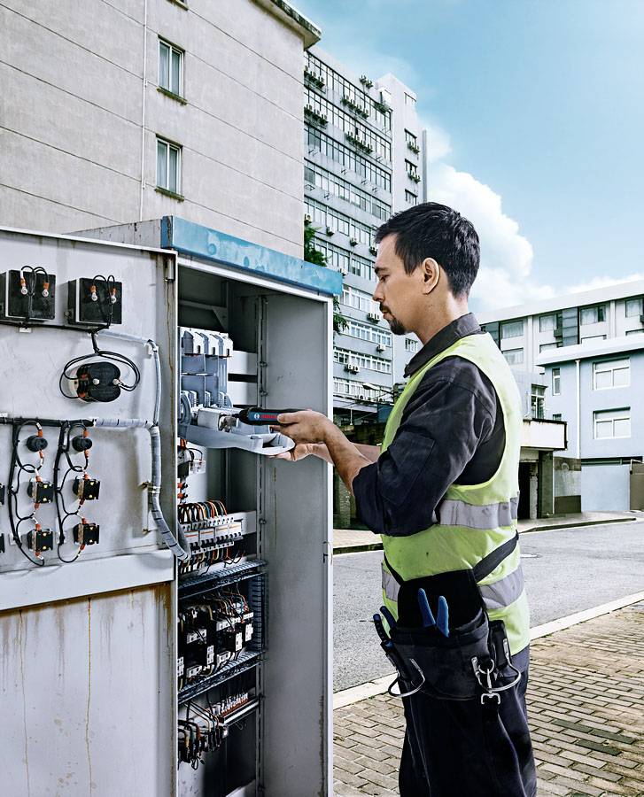 Un technicien en tenue de travail et gilet de sécurité répare un coffret de distribution électrique ouvert sur une rue urbaine.